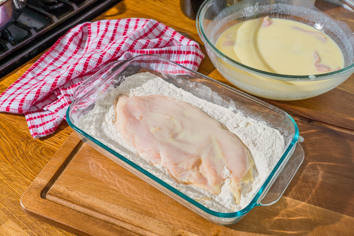 A glass baking dish with seasoned flour and a piece of buttermilk soaked chicken on top. The dish is on a wooden counter.