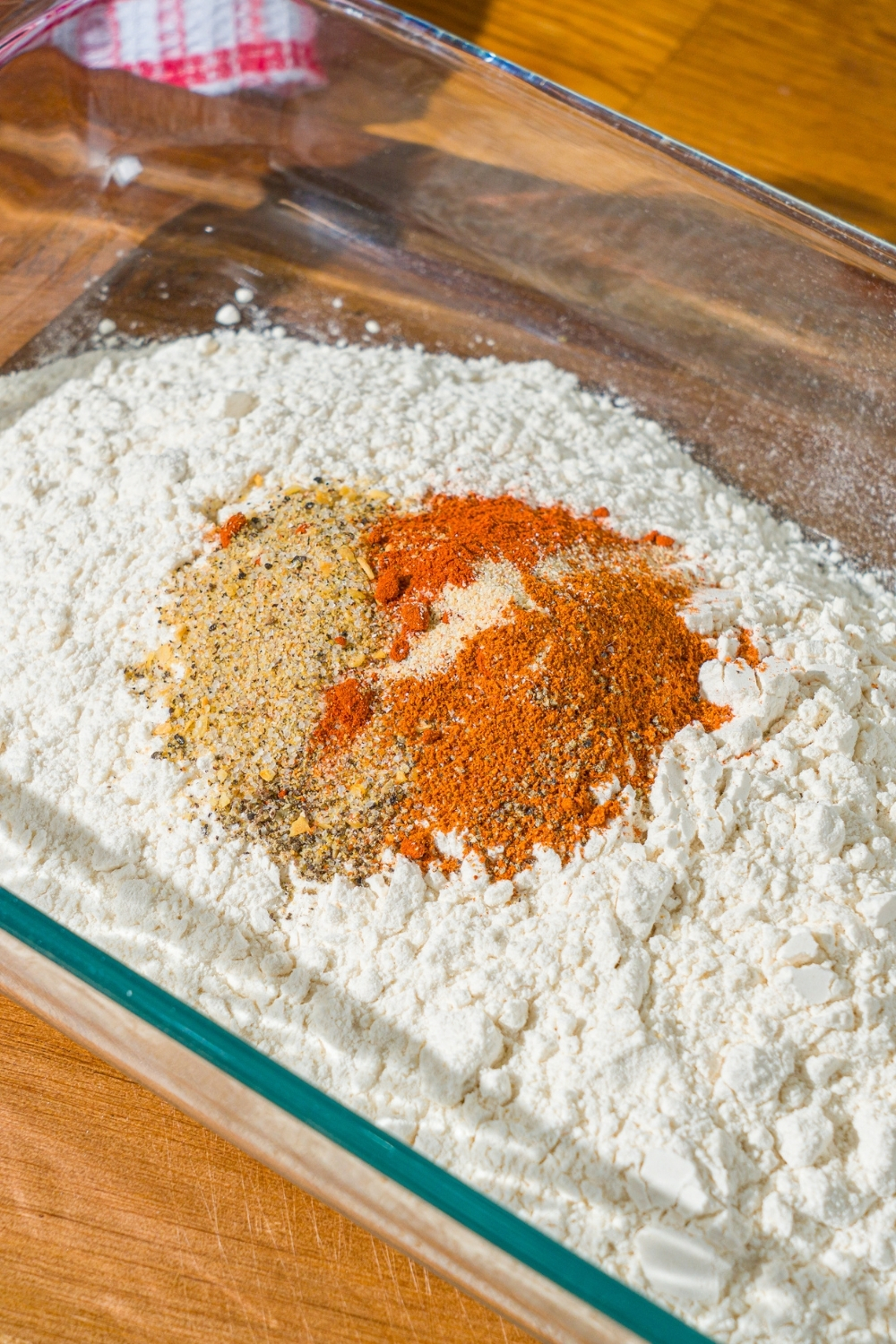A glass baking dish with flour topped with seasonings including breadcrumbs, paprika, cayenne pepper, and more. The dish is on a wooden counter.