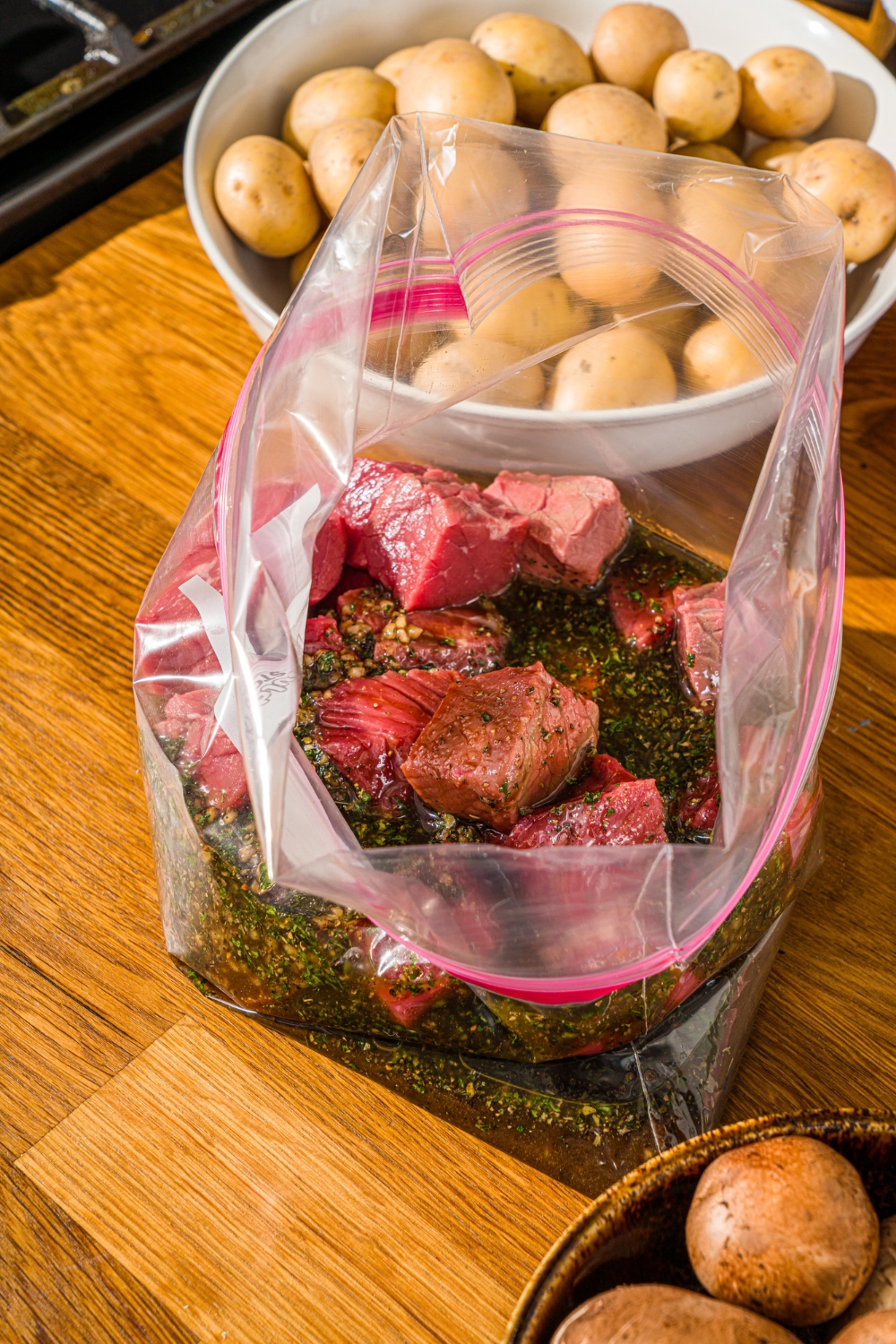 A large ziploc bag with steak marinating in a kabob marinade. The bag is on a wooden counter with a bowl of potatoes.