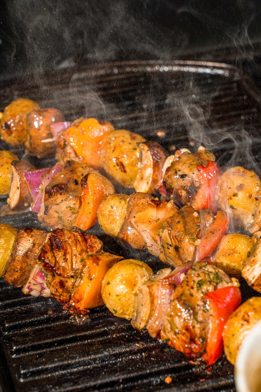Three steak kabobs with marinated steak, peppers, onions, and potatoes grilling on a grill.