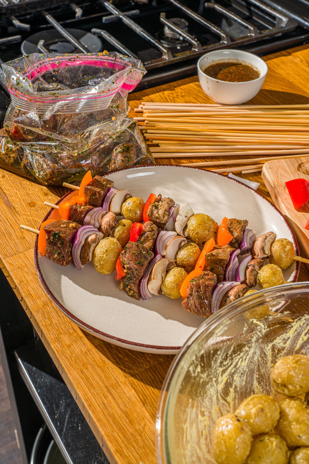 A white plate with three uncooked steak kabobs with marinated steak, peppers, onions, and potatoes. The plate is on a wooden counter with a bag of marinade and bowl of potatoes.