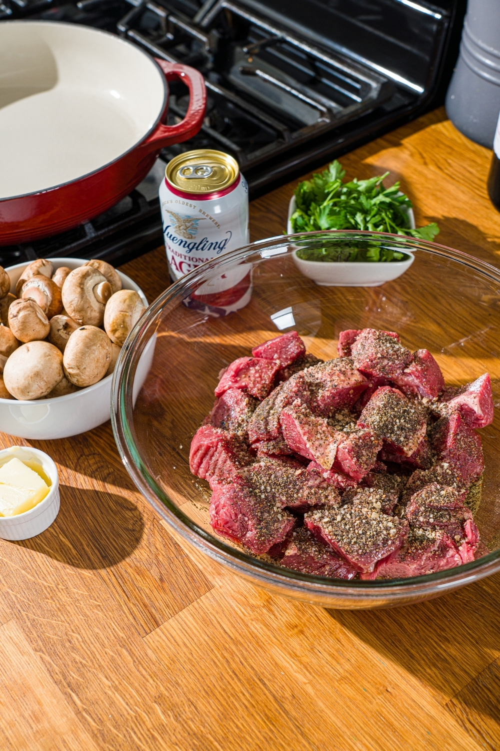 A glass bowl with uncooked beef tips topped with seasonings. The bowl is on a wooden counter with additional ingredients.
