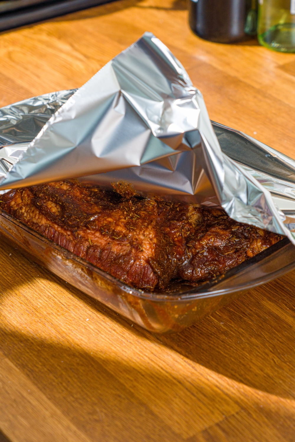 A glass baking dish with uncooked beef brisket rubbed with bbq sauce and seasonings. A piece of foil is covering the dish.