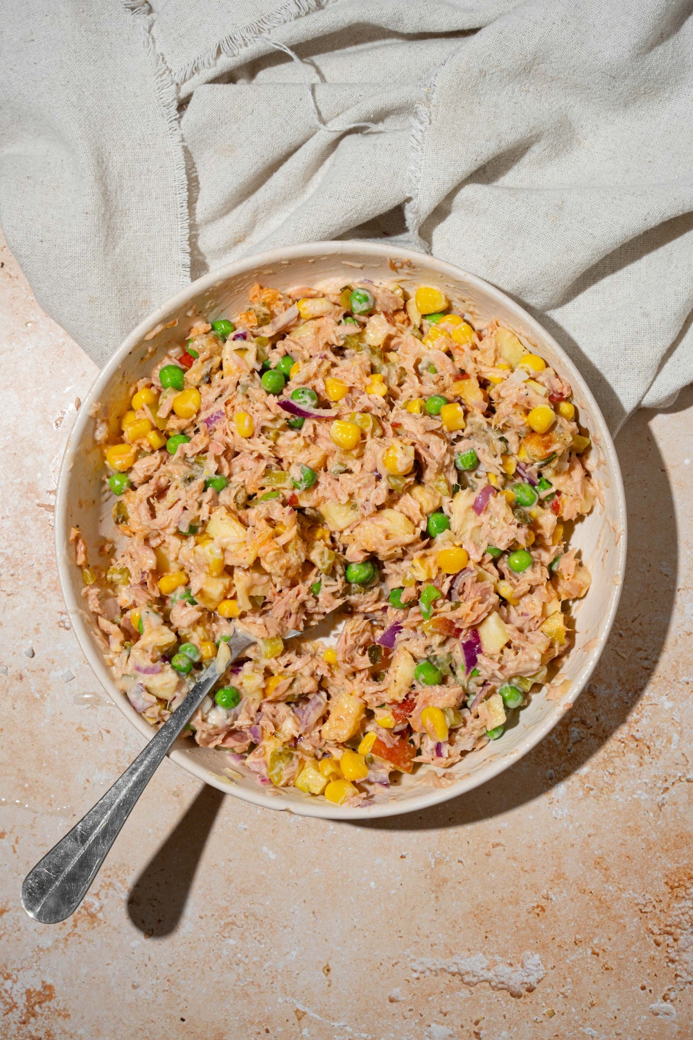 A large white bowl with a spoon mixing ingredients to make Matthew McConaughey tuna salad including tuna, peas, corn, apple, jalapeño chips, pickles, and red onion tossed in a wasabi mayonnaise dressing. The bowl is on a tan counter with a white cloth napkin.