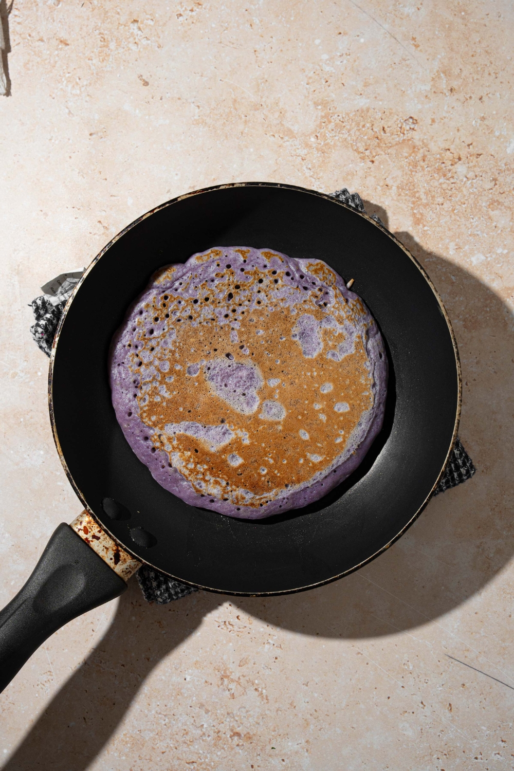 A skillet with a flipped ube pancake. The skillet is on a dish rag on a tan counter.