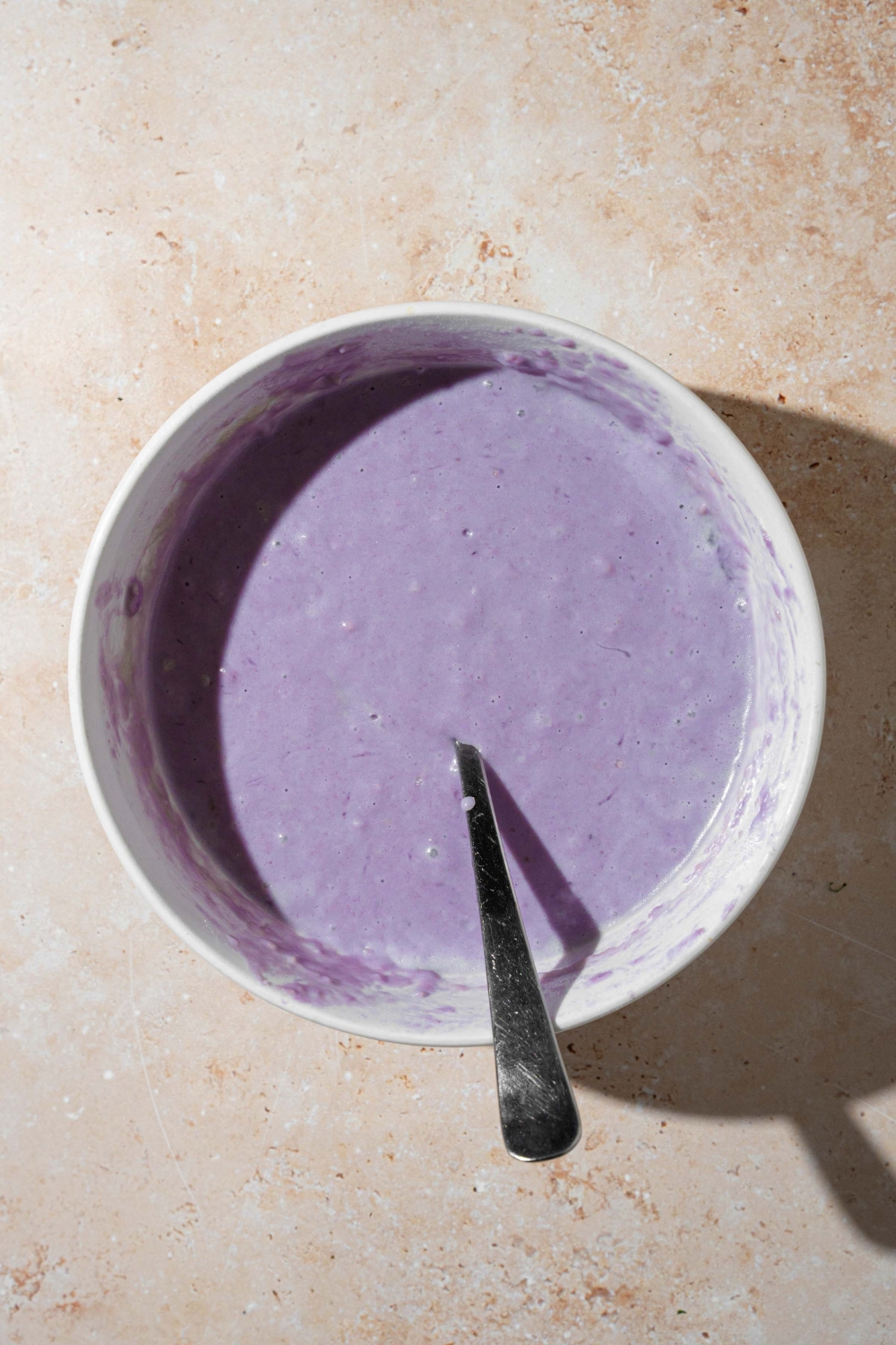 A large white bowl with a spoon in ube pancake batter. The bowl is on a tan counter.