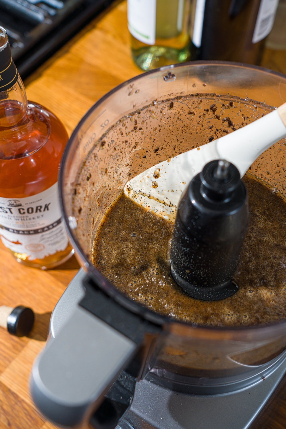 A food processor with blended vanilla bean pods, seeds, corn syrup, and whiskey. A spatula is scraping the sides of the food processor.