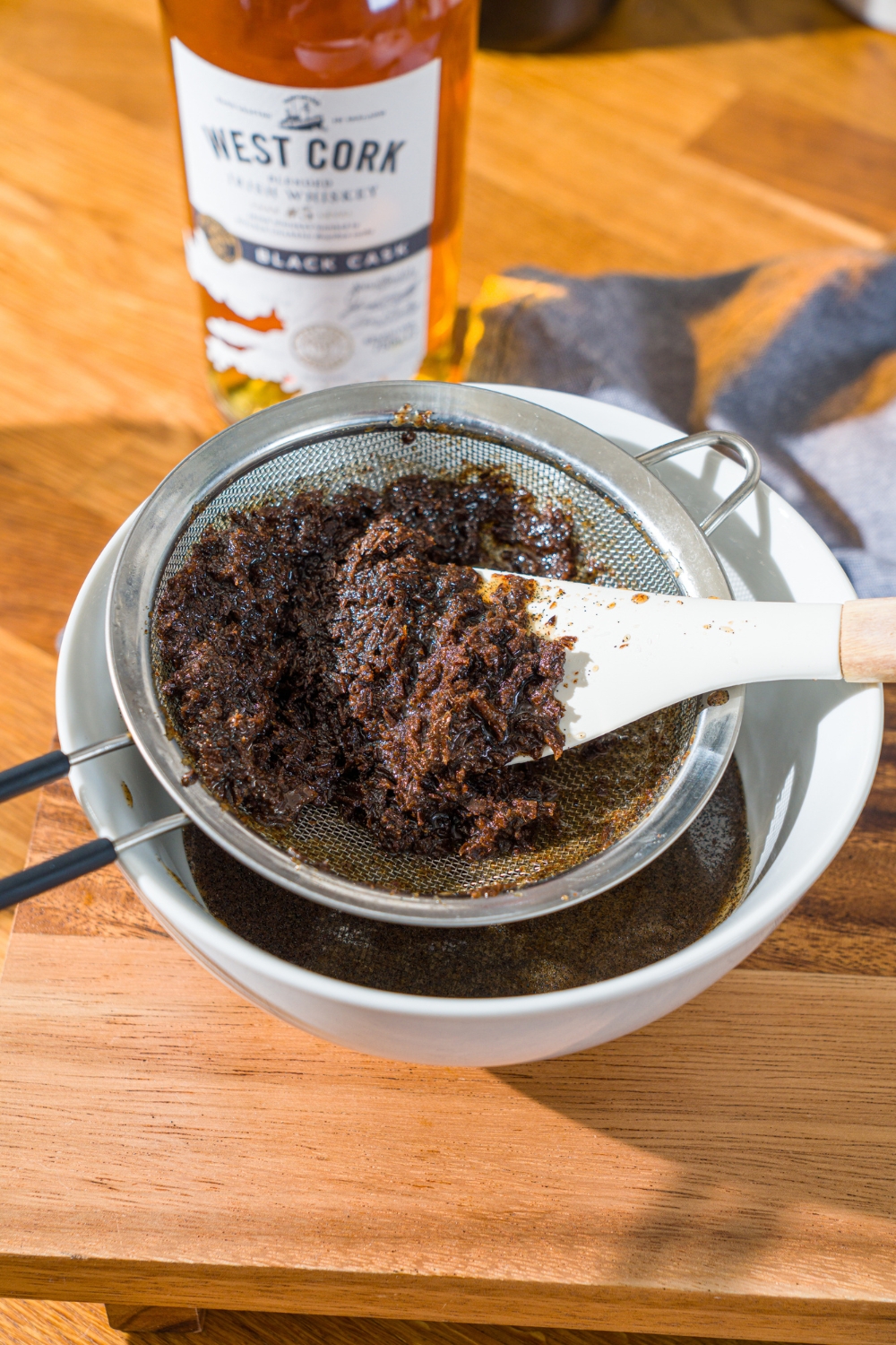 A vanilla bean paste mixture straining through a mesh strainer over a white bowl. A spatula is collecting the seeds strained from the mixture.