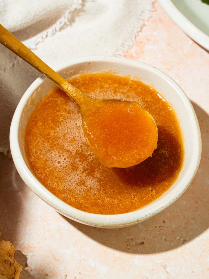 A small white bowl of hibachi ginger sauce with a spoon in the bowl. The bowl is on a tan counter with ginger, a plate with lettuce, and a white cloth napkin.