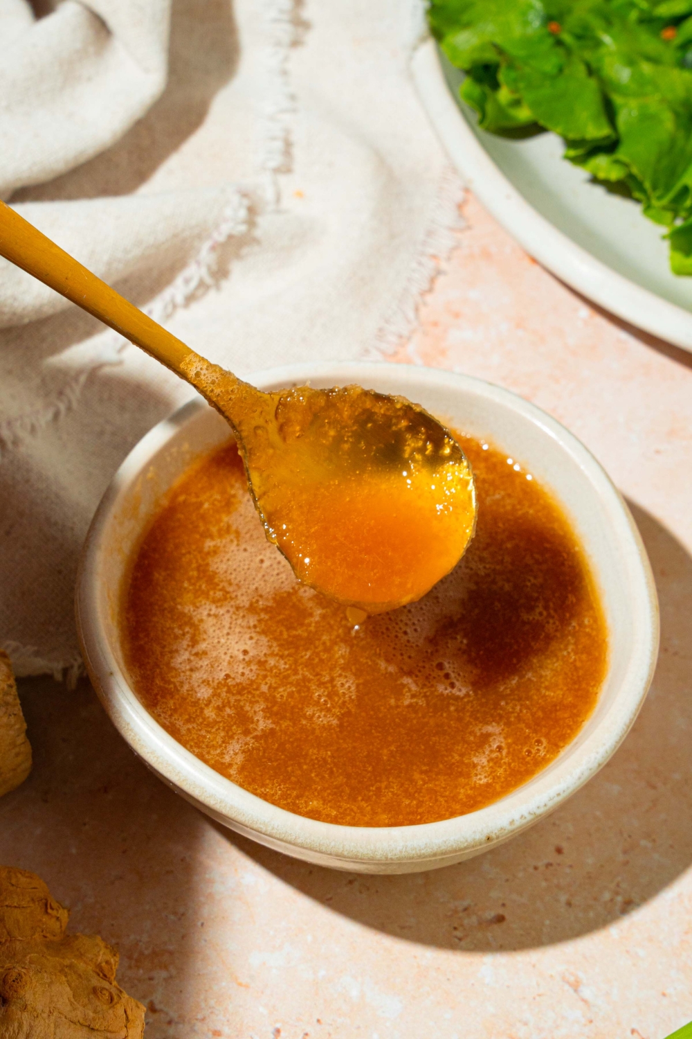A small white bowl of hibachi ginger sauce with a spoon dipping in the bowl. The bowl is on a tan counter with ginger, a plate with lettuce, and a white cloth napkin.