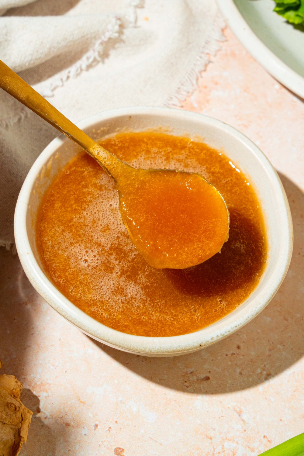 A small white bowl of hibachi ginger sauce with a spoon in the bowl. The bowl is on a tan counter with ginger, a plate with lettuce, and a white cloth napkin.
