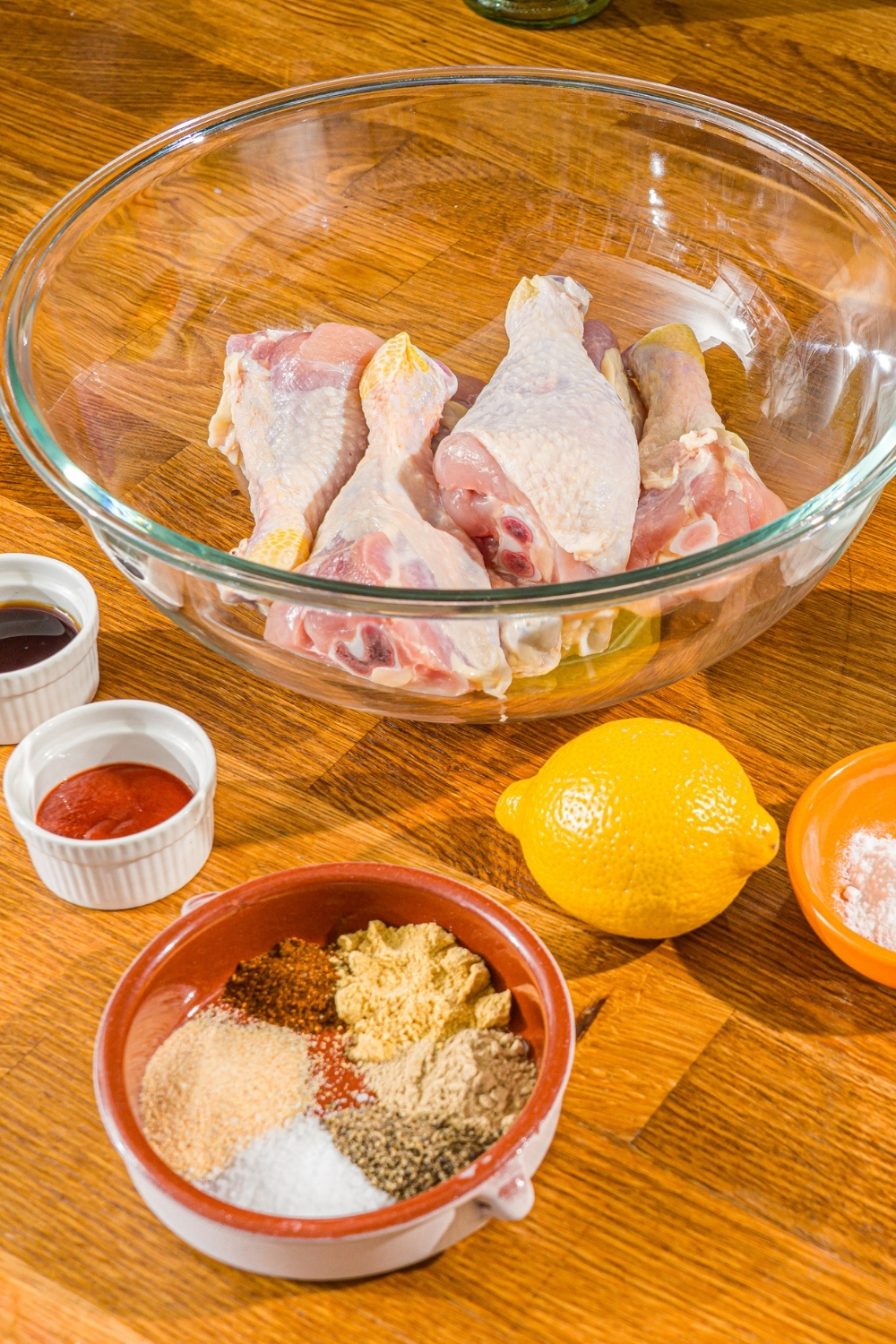 A wooden counter with ingredients to make copycat KFC fried chicken including a bowl of chicken legs, lemon, and a bowl of seasonings.