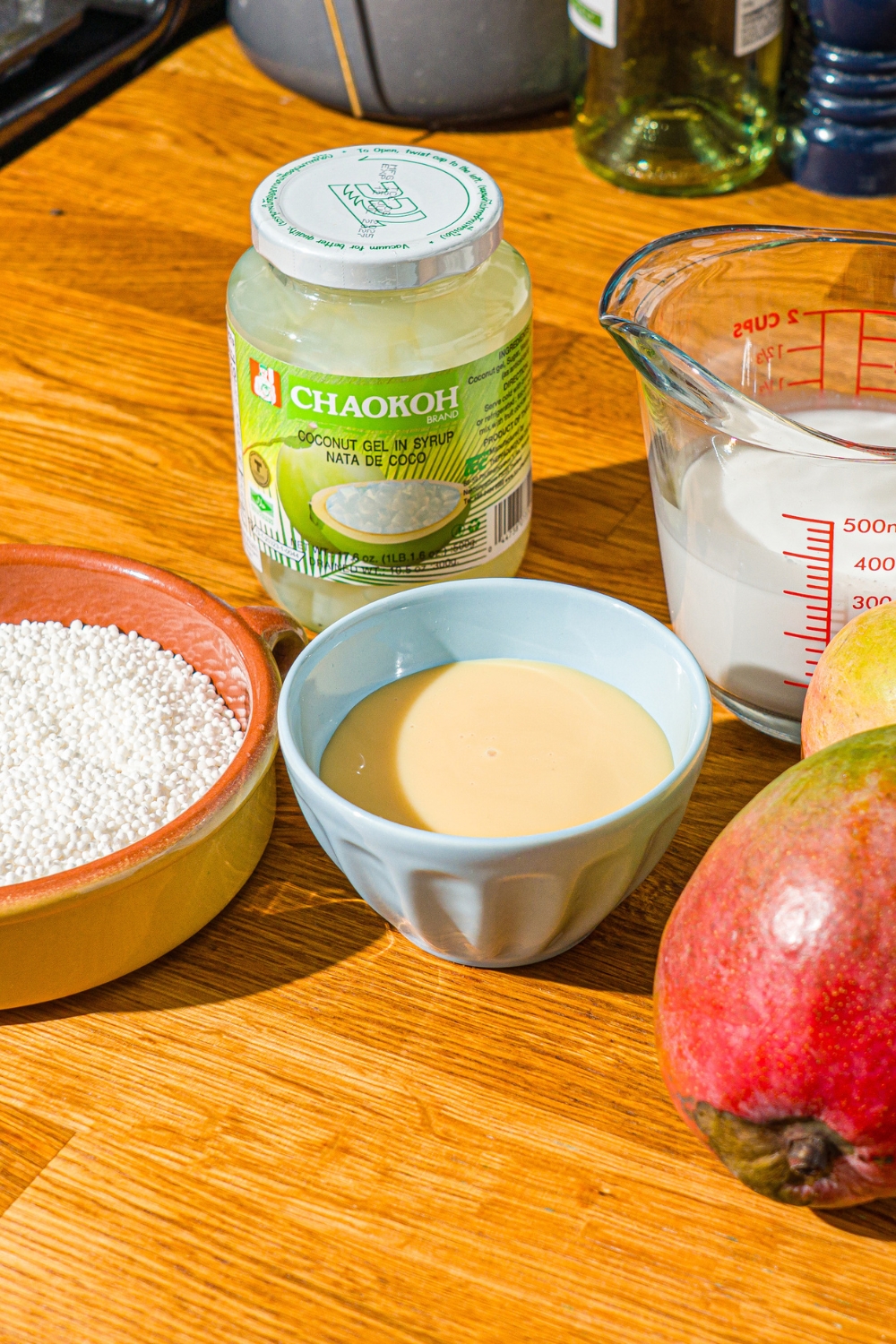 A wooden counter with ingredients to make mango sago including sago pearls, coconut jelly, mango, condensed milk, and coconut milk.