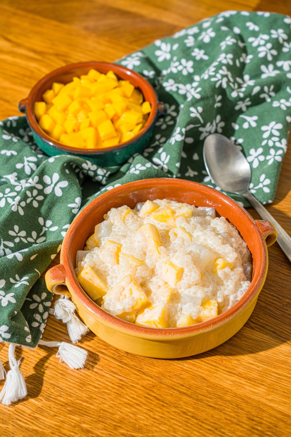A small bowl of mango sago topped with diced mango. The bowl is on a wooden counter with a spoon, green floral napkin, and small bowl of diced mango.