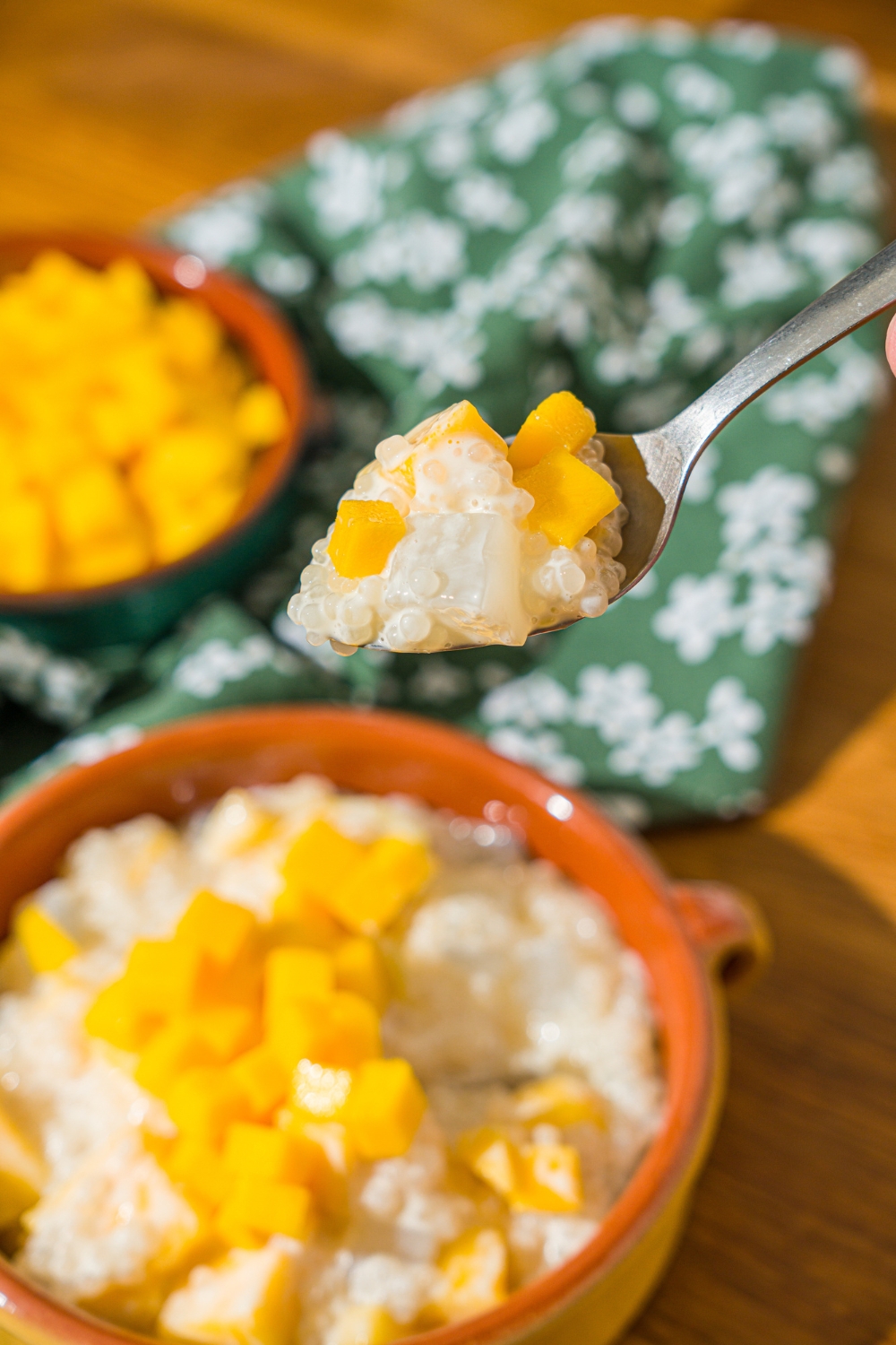 A close up of a spoon with a bite of mango sago. There is a bowl of mango sago on a wooden counter with a green floral napkin.