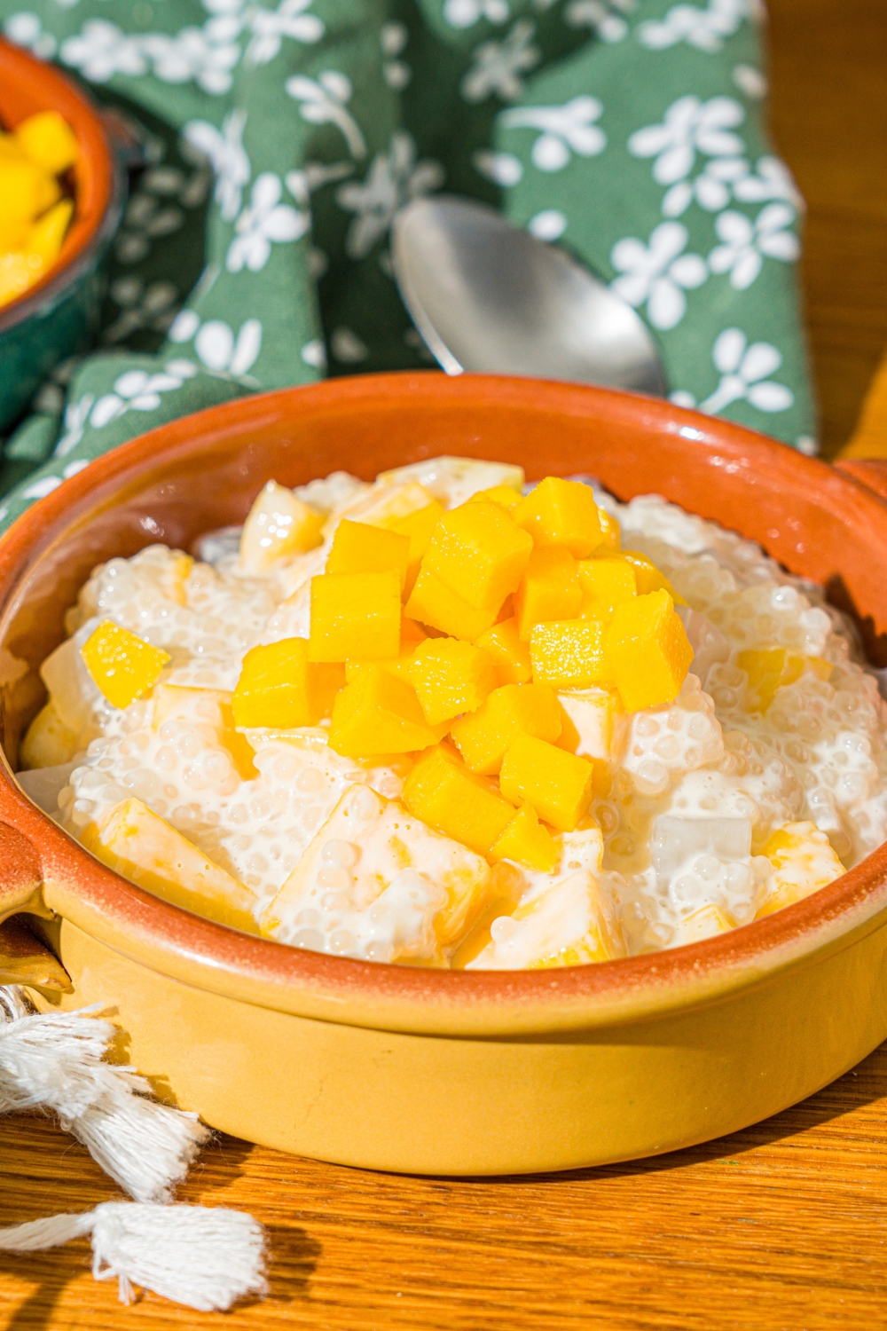 A small bowl of mango sago topped with diced mango. The bowl is on a wooden counter with a spoon, green floral napkin, and small bowl of diced mango.