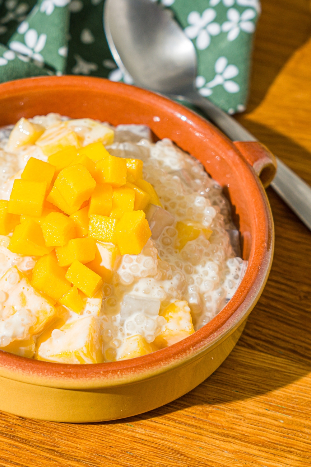 A small bowl of mango sago topped with diced mango. The bowl is on a wooden counter with a spoon, and green floral napkin.