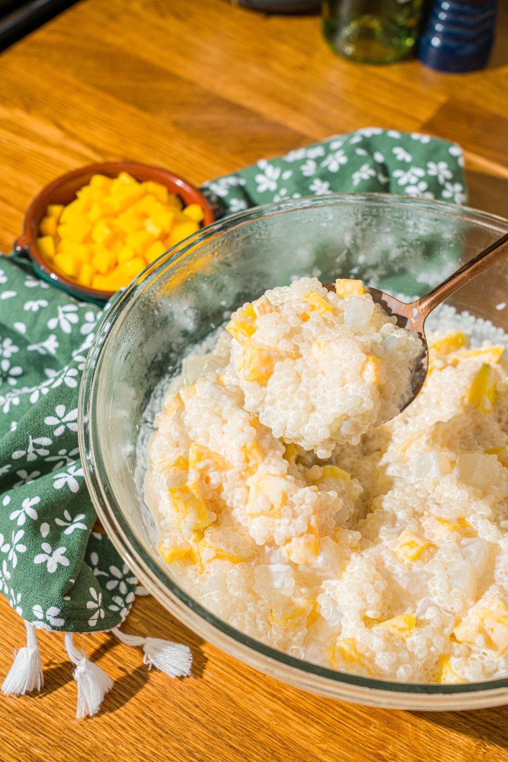 A glass bowl with a mango sago mixture. A spoon is scooping a bite of mango sago from the bowl. The bowl is on a wooden counter with a green floral napkin and small bowl of diced mango.