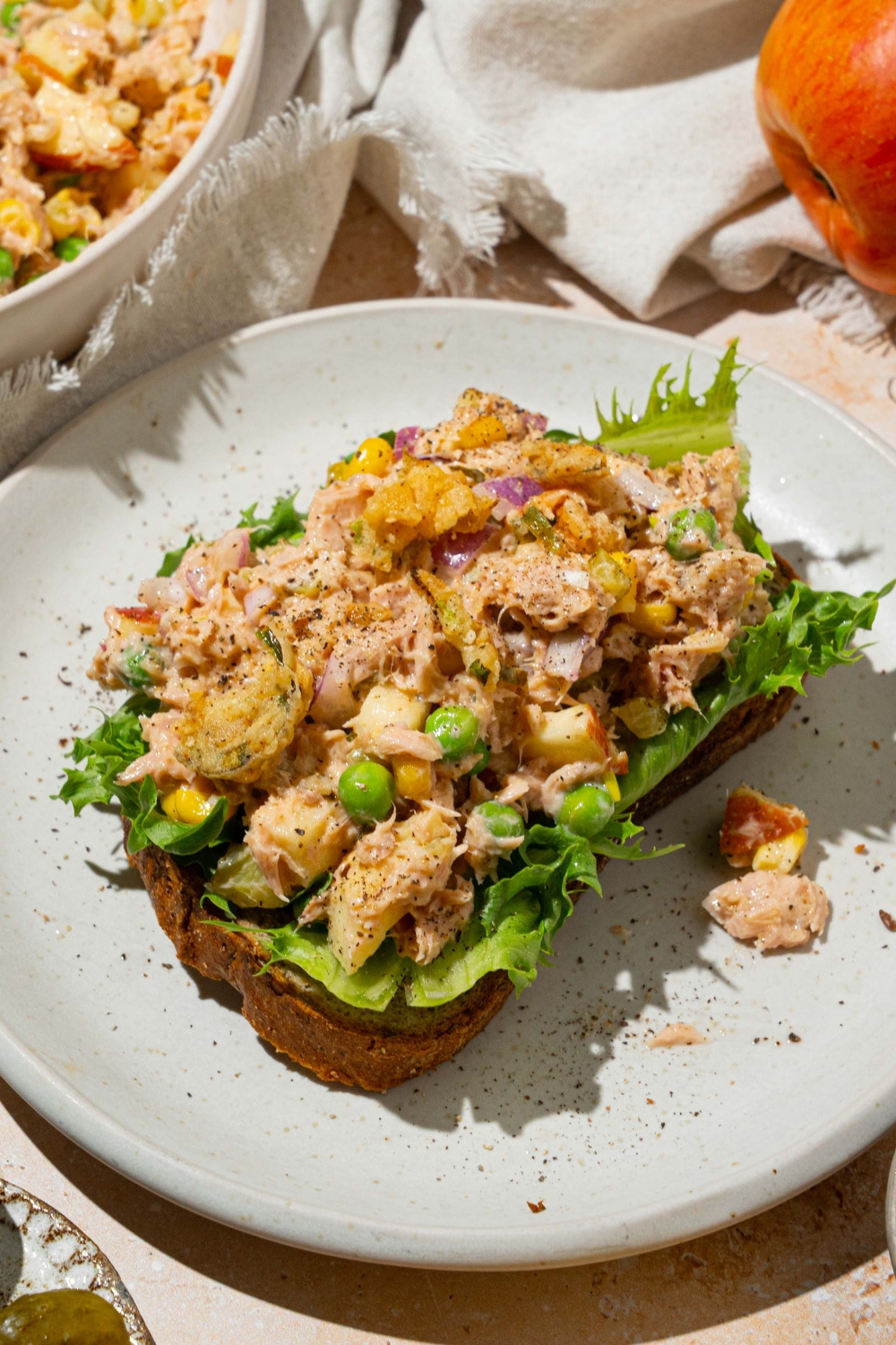 A white plate with a slice of bread topped with lettuce and spread with Matthew McConaughey tuna salad including tuna, peas, corn, apple, pickles, red onion, and jalapeño chips in a wasabi mayonnaise dressing. The plate is on a tan counter with a white cloth napkin and a bowl of tuna.