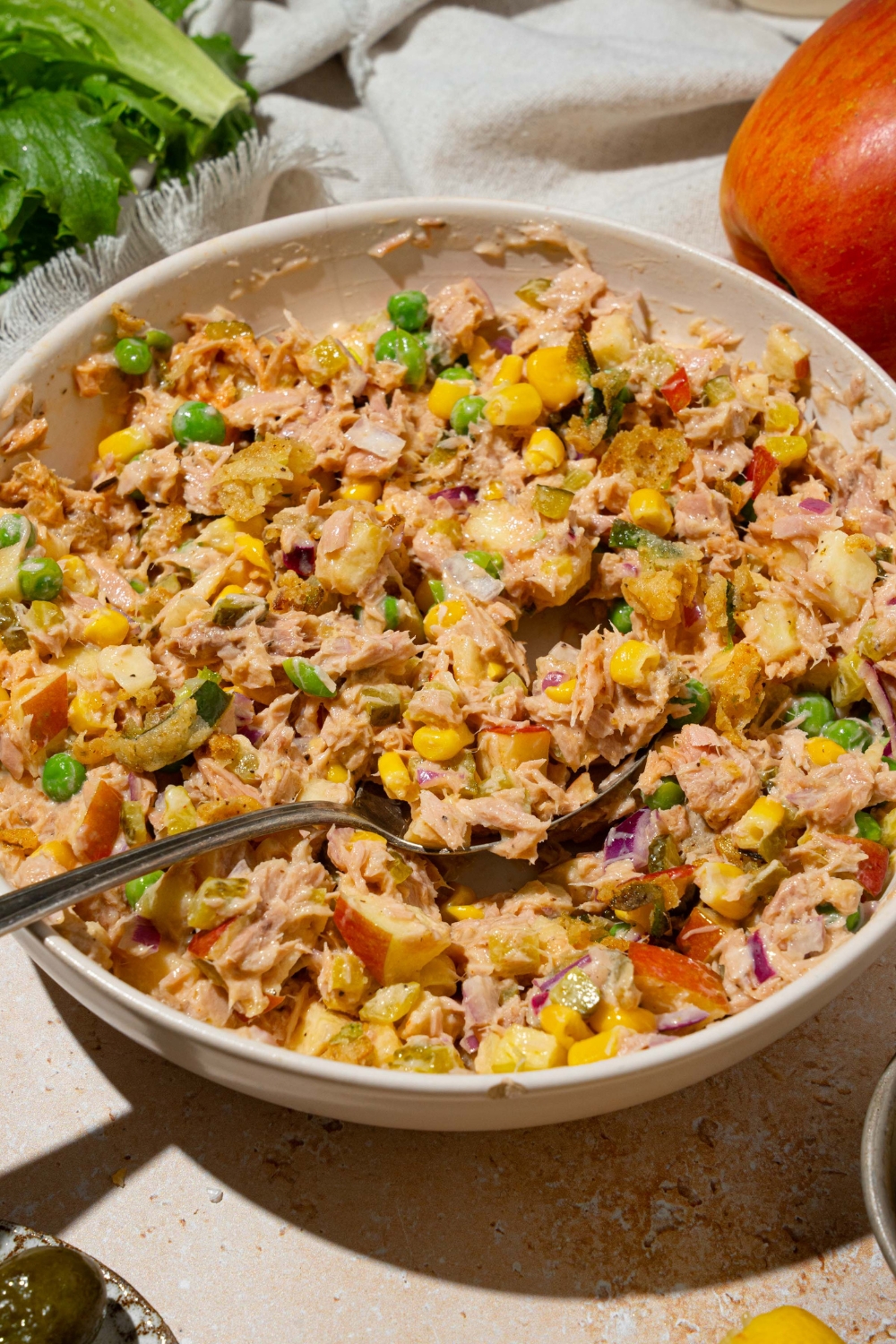 A large white bowl with Matthew McConaughey tuna salad including tuna, corn, peas, apple, pickles, red onion, and jalapeño chips in a wasabi mayo dressing. There is a spoon in the bowl. The bowl is on a tan counter with a white cloth napkin.