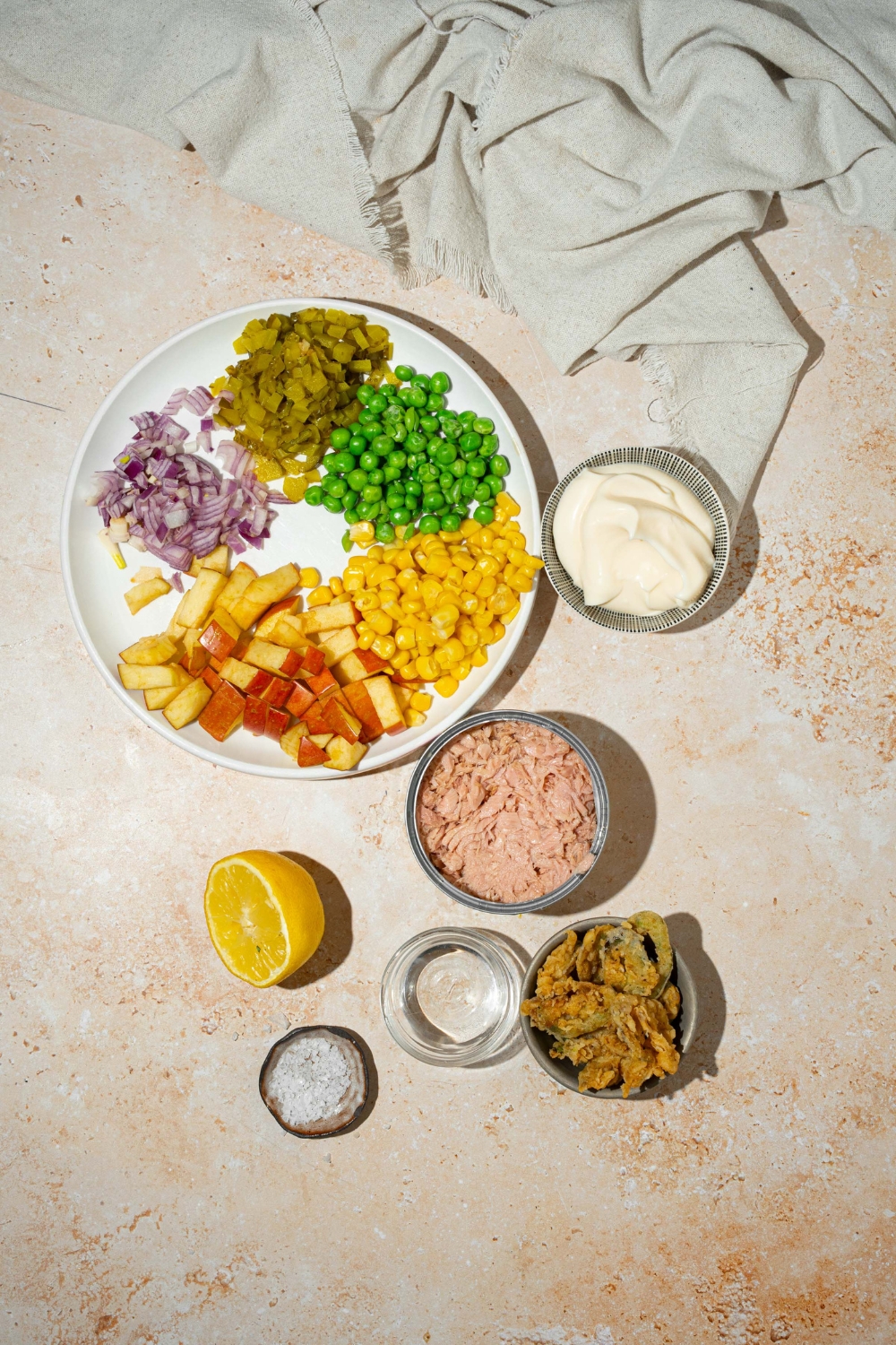 A tan counter with several bowls containing ingredients to make Matthew McConaughey tuna salad including tuna, corn, peas, apple, pickles, jalapeño chips, wasabi paste, mayo, vinegar, and lemon.