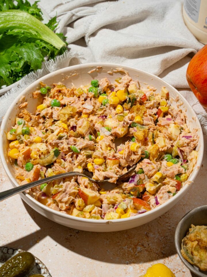 A large white bowl with Matthew McConaughey tuna salad including tuna, corn, peas, apple, pickles, red onion, and jalapeño chips in a wasabi mayo dressing. There is a spoon in the bowl. The bowl is on a tan counter with a white cloth napkin.