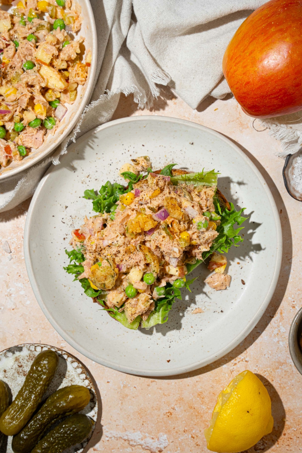 A white plate with a slice of bread topped with lettuce and spread with Matthew McConaughey tuna salad including tuna, peas, corn, apple, pickles, red onion, and jalapeño chips in a wasabi mayonnaise dressing. The plate is on a tan counter with a white cloth napkin and a bowl of tuna.