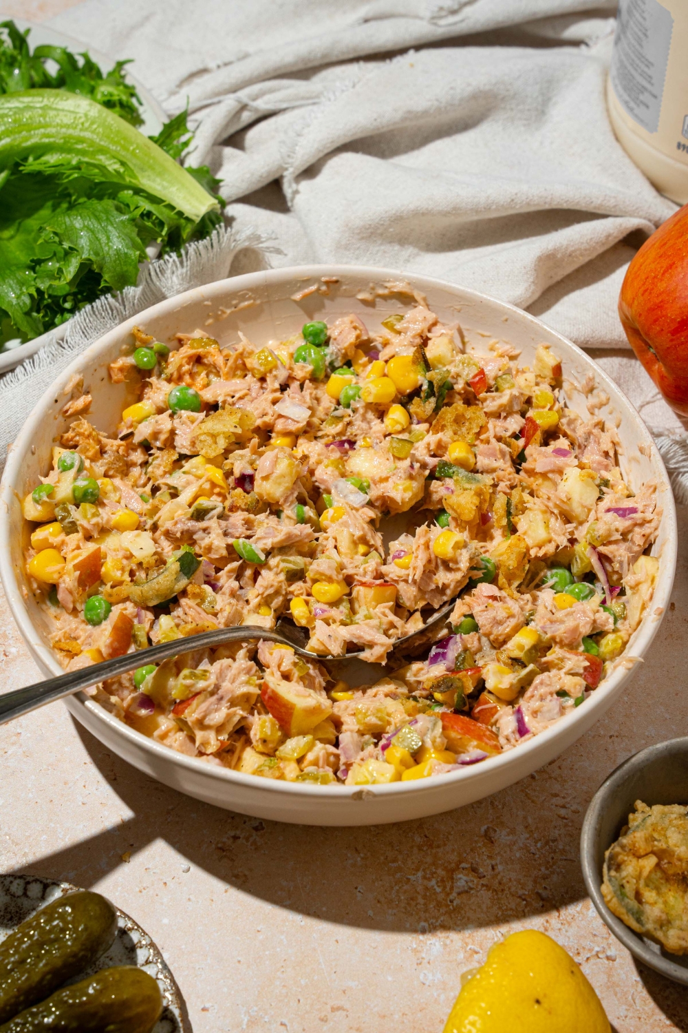 A large white bowl with Matthew McConaughey tuna salad including tuna, corn, peas, apple, pickles, red onion, and jalapeño chips in a wasabi mayo dressing. There is a spoon in the bowl. The bowl is on a tan counter with a white cloth napkin.