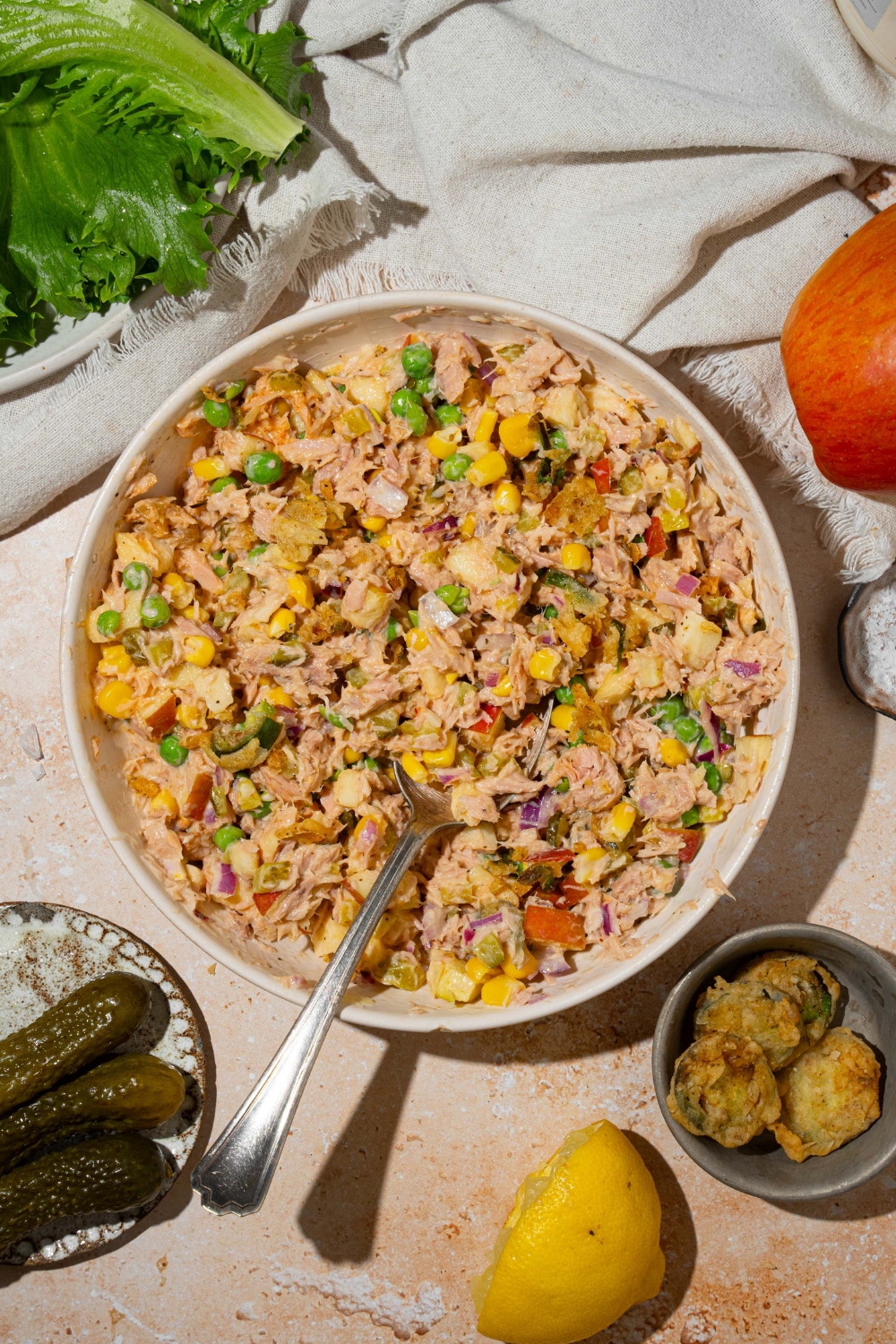A large white bowl with Matthew McConaughey tuna salad including tuna, corn, peas, apple, pickles, red onion, and jalapeño chips in a wasabi mayo dressing. There is a spoon in the bowl. The bowl is on a tan counter with a white cloth napkin.