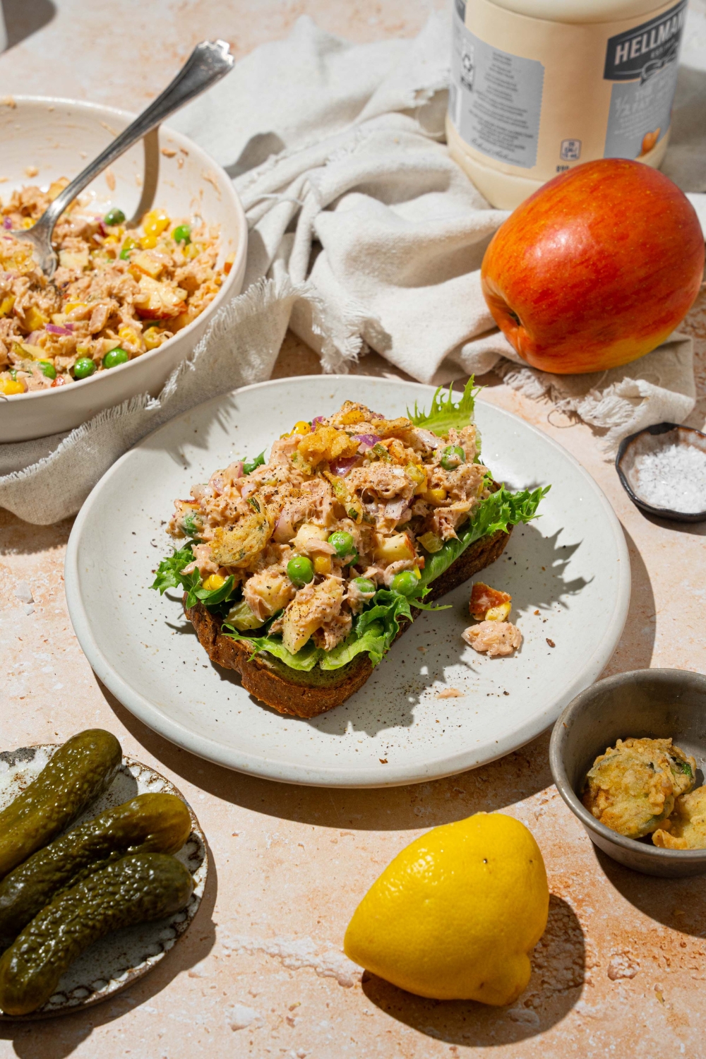 A white plate with a slice of bread topped with lettuce and spread with Matthew McConaughey tuna salad including tuna, peas, corn, apple, pickles, red onion, and jalapeño chips in a wasabi mayonnaise dressing. The plate is on a tan counter with a white cloth napkin and a bowl of tuna.