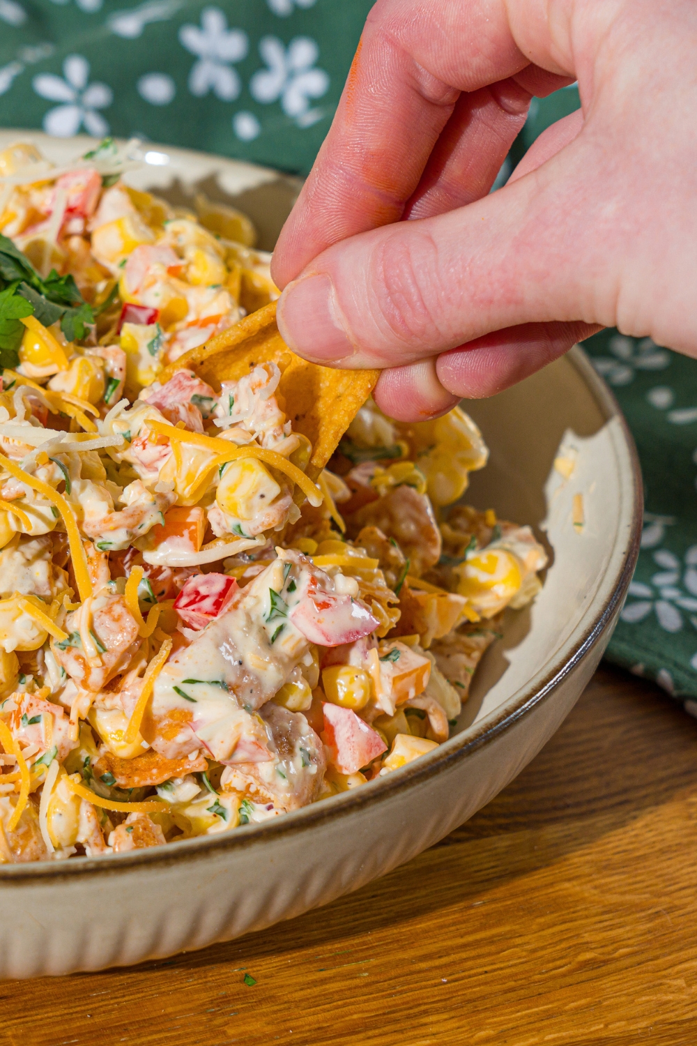 A white bowl with corn salad with chili cheese fritos garnished with fresh parsley. A hand is dipping a chip into the bowl. The bowl is on a wooden counter with a green floral napkin.