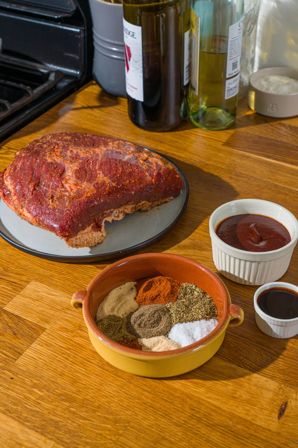 A wooden counter with ingredients to make oven baked beef brisket including brisket, bbq sauce, and seasonings.