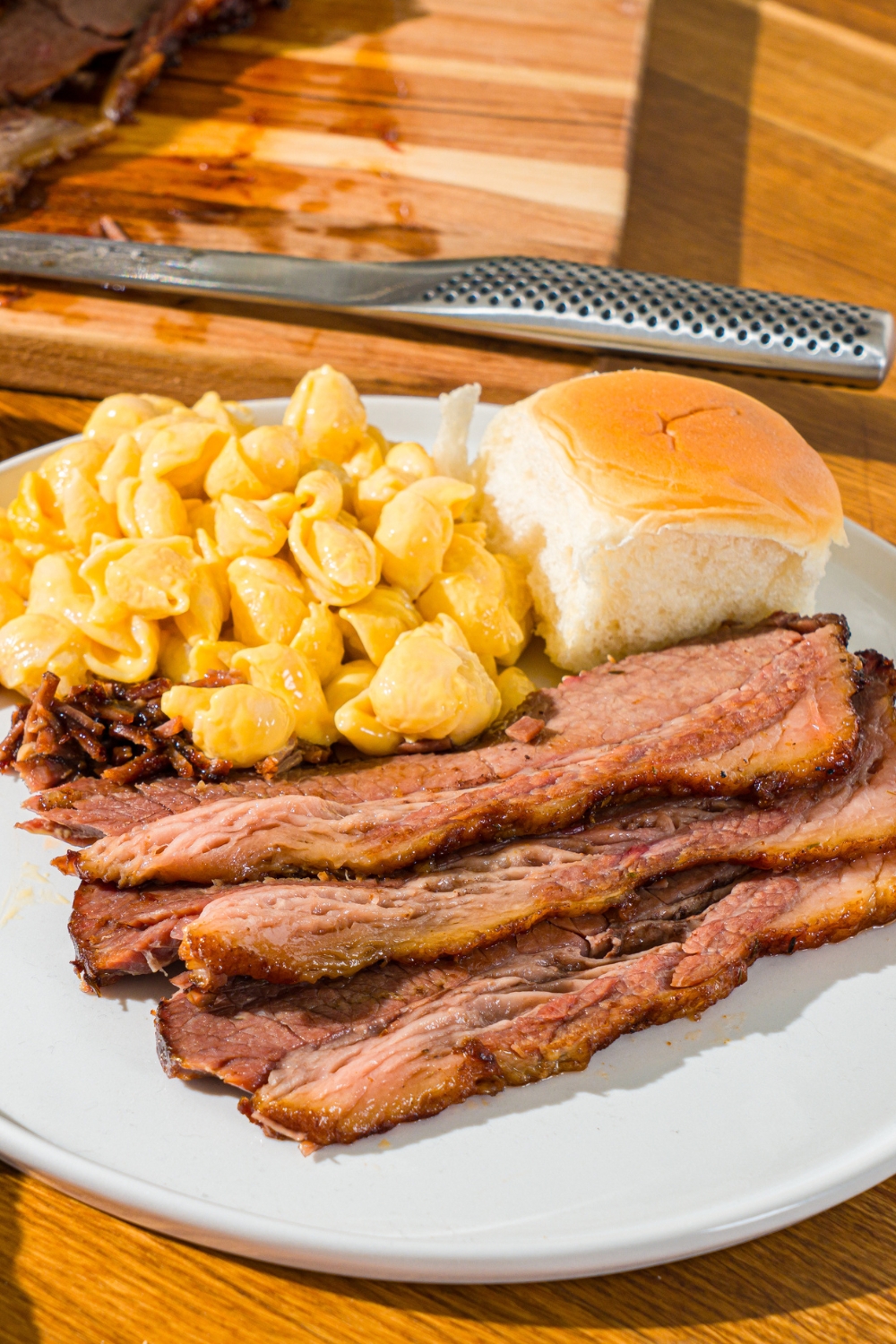 A white plate with sliced oven baked beef brisket, mac and cheese, and a dinner roll. The plate is on a wooden counter.
