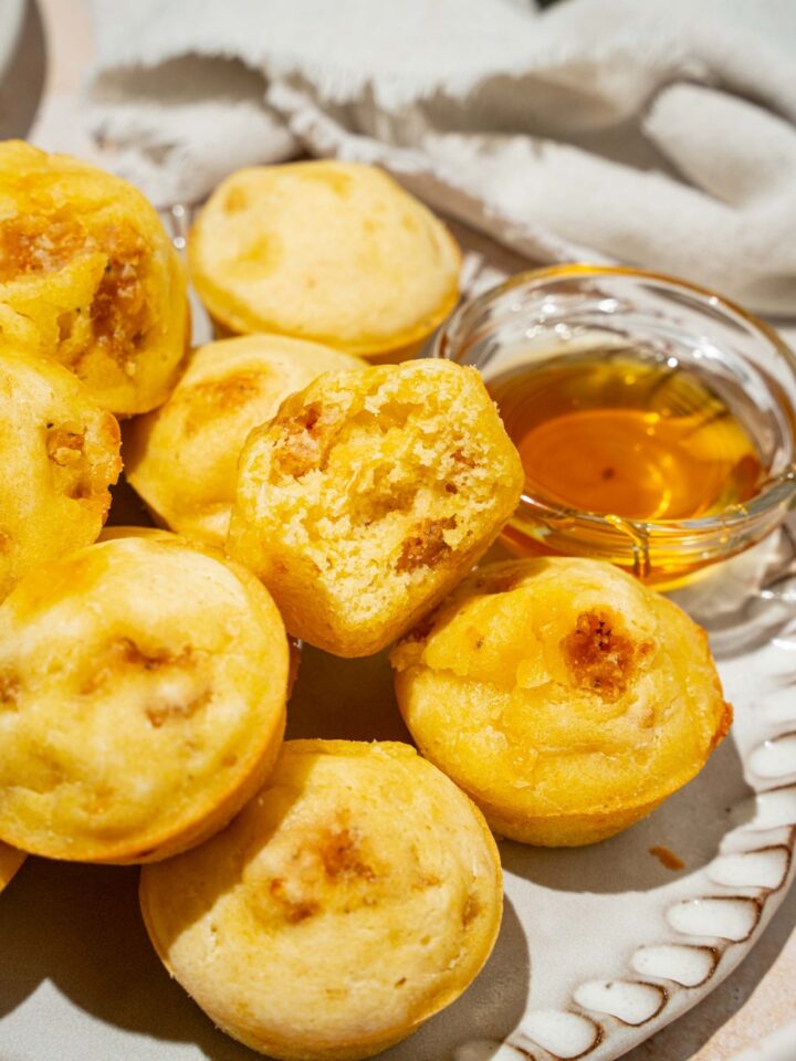 A plate with a pile of pancake sausage muffins and small bowl of syrup. There is a bite taken from one of the muffins. The plate is on a tan counter.