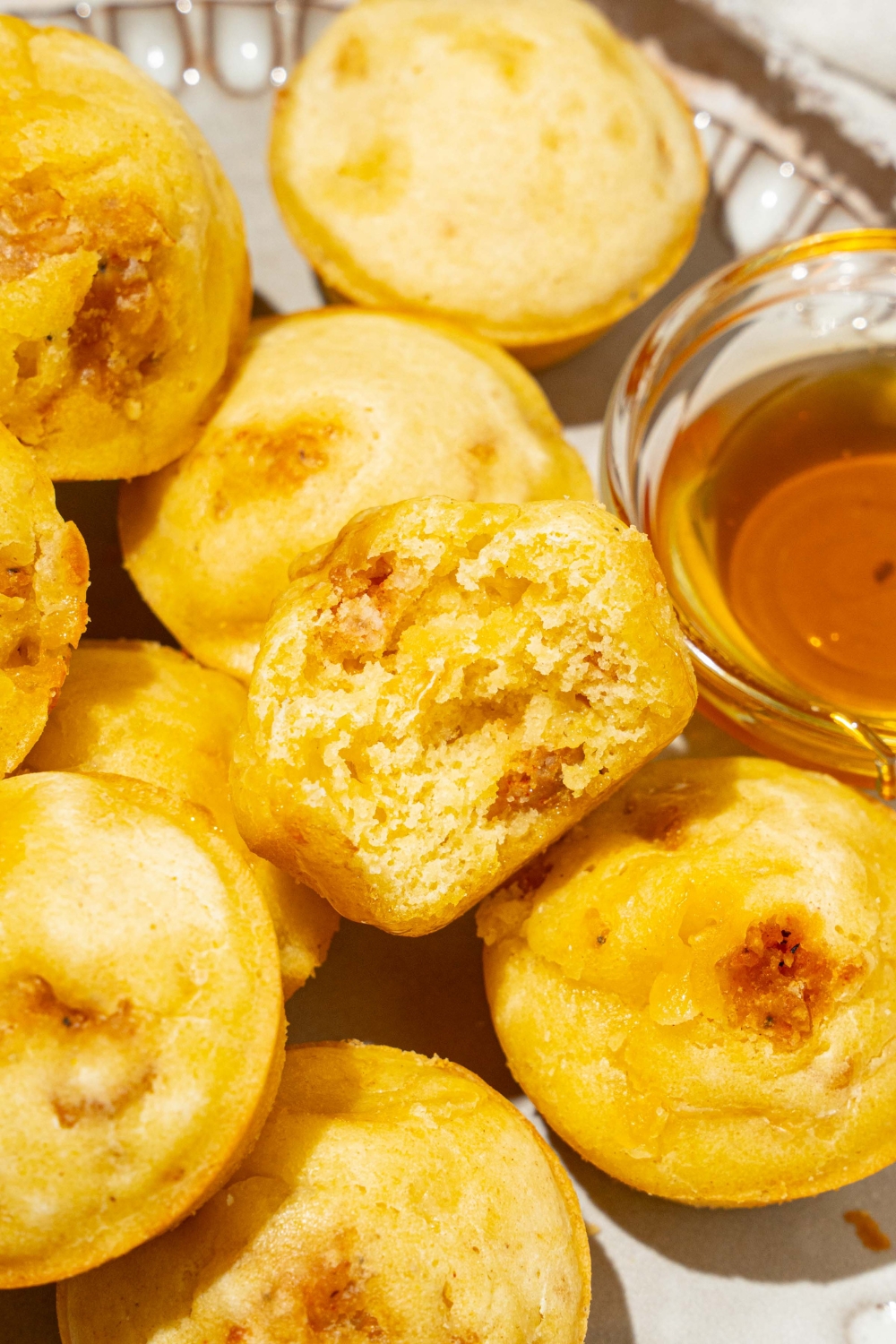 A plate with a pile of pancake sausage muffins and small bowl of syrup. There is a bite taken from one of the muffins. The plate is on a tan counter.