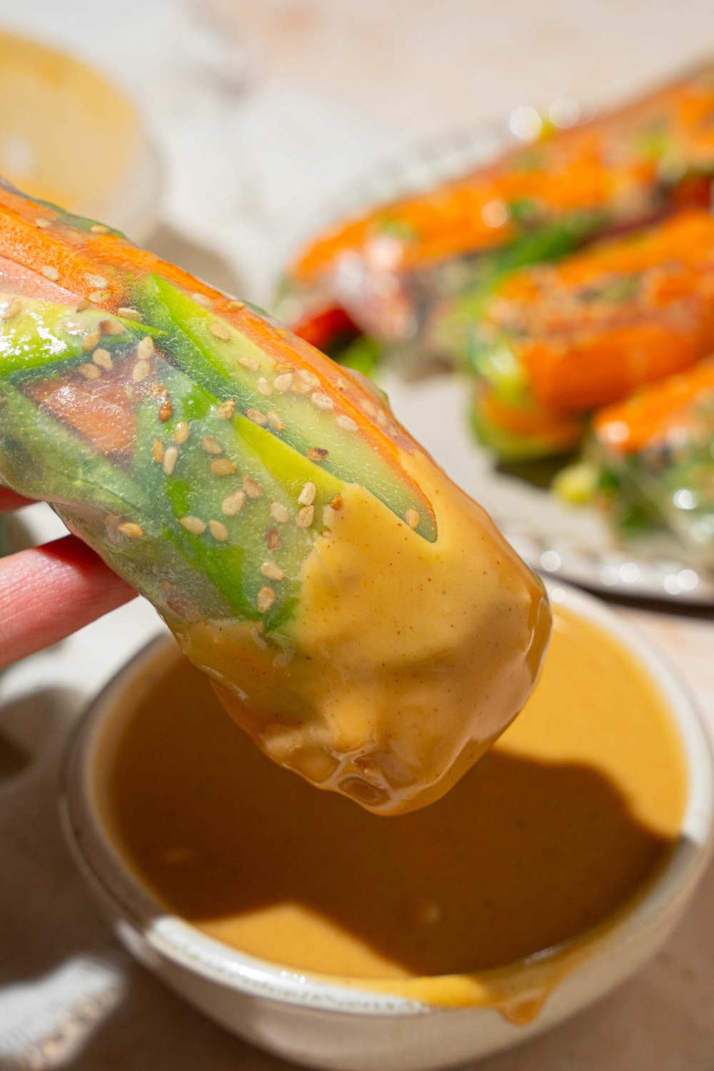 A fresh spring roll dipping into a small white bowl of peanut sauce. The bowl is on a tan counter with a plate of spring rolls.