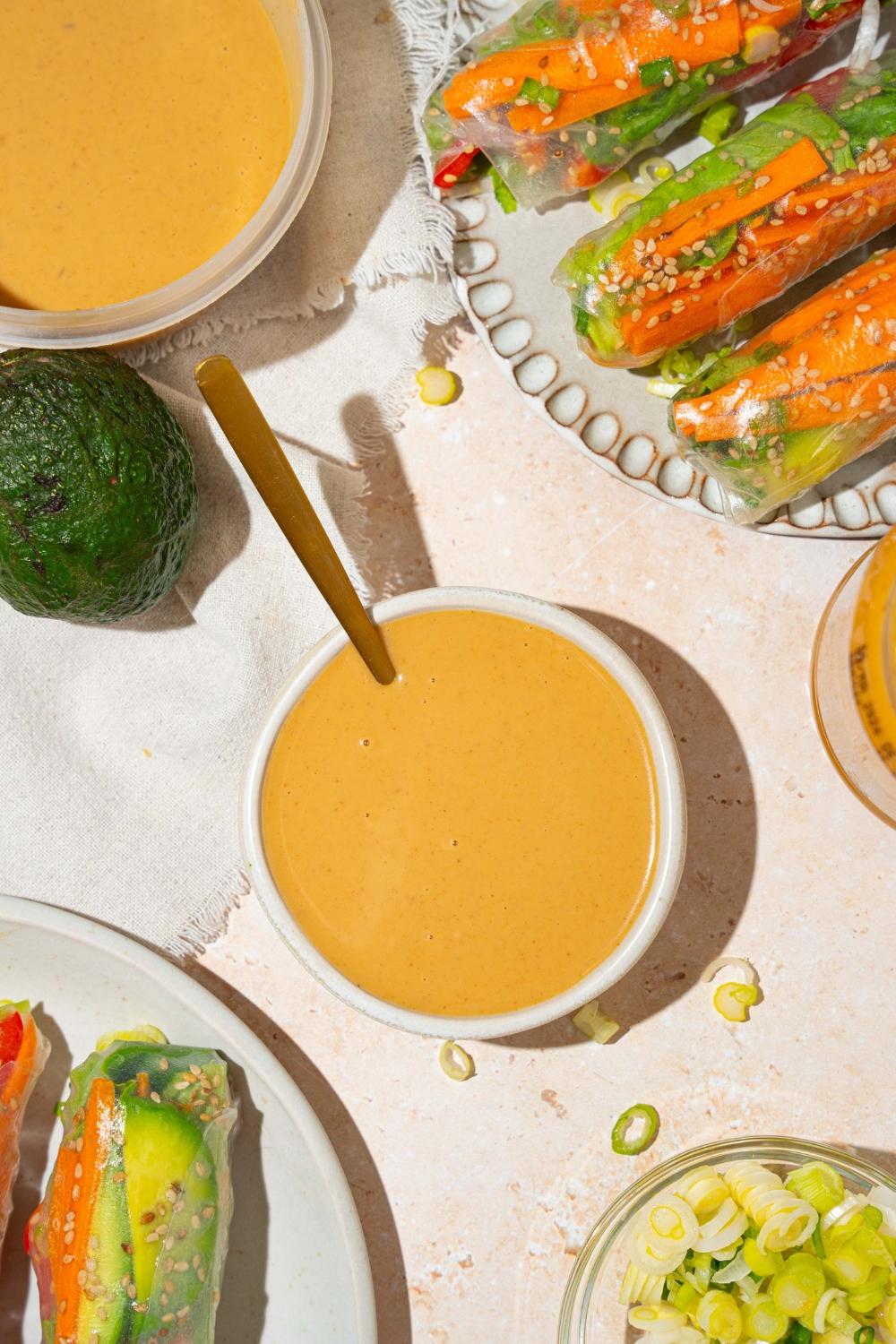 A small white bowl of peanut sauce with a spoon in the bowl. The bowl is on a tan counter with a plate of spring rolls and an additional bowl of sauce.