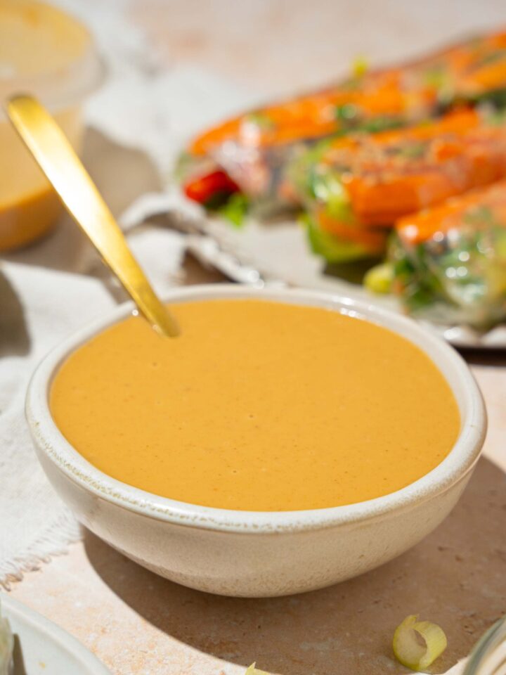 A small white bowl of peanut sauce with a spoon in the bowl. The bowl is on a tan counter with a plate of spring rolls and an additional bowl of sauce.