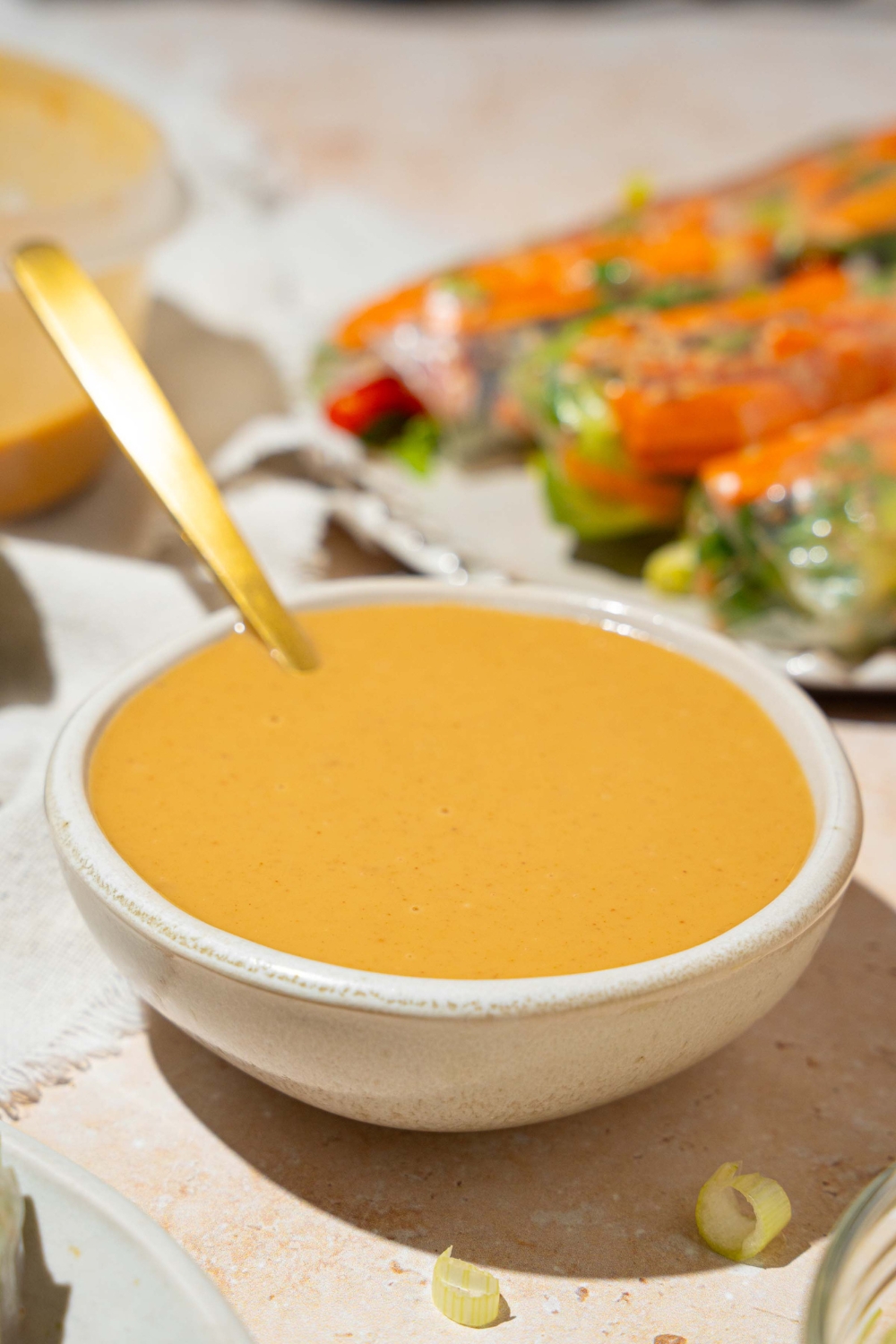 A small white bowl of peanut sauce with a spoon in the bowl. The bowl is on a tan counter with a plate of spring rolls and an additional bowl of sauce.
