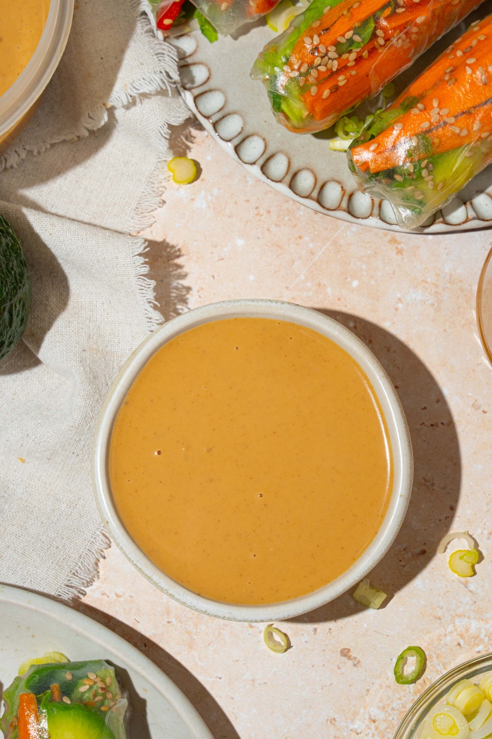 A small white bowl of peanut sauce. The bowl is on a tan counter with a plate of spring rolls and an additional bowl of sauce.