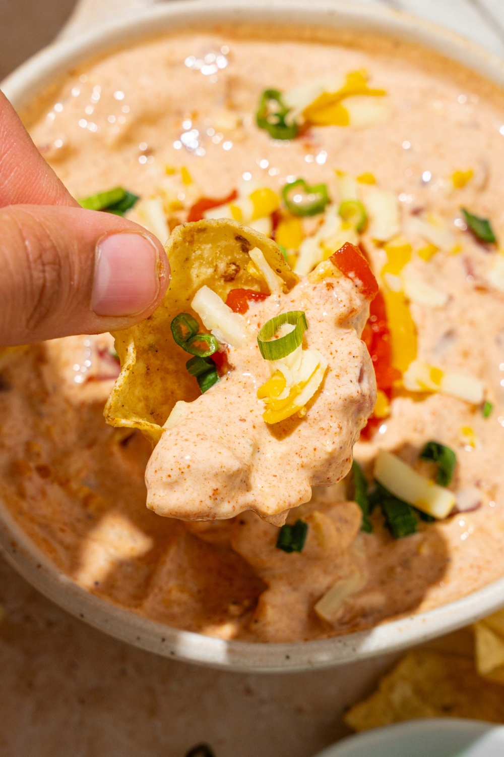A white bowl with poolside dip garnished with sliced green onion and shredded cheese. A chip is being dipped into the bowl.