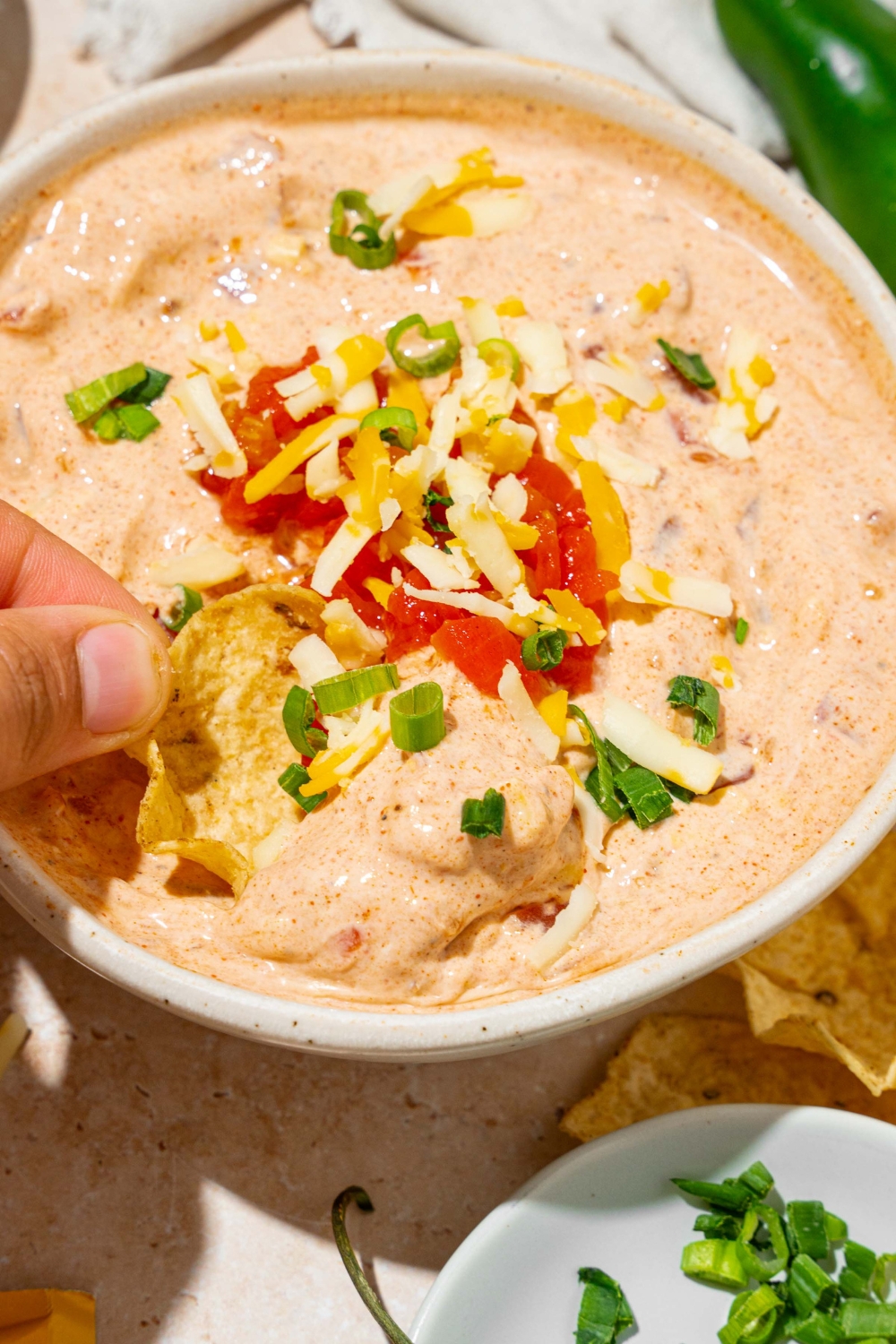 A white bowl with poolside dip garnished with sliced green onion and shredded cheese. A chip is being dipped into the bowl. The bowl is on a tan counter with a small plate of garnishes.