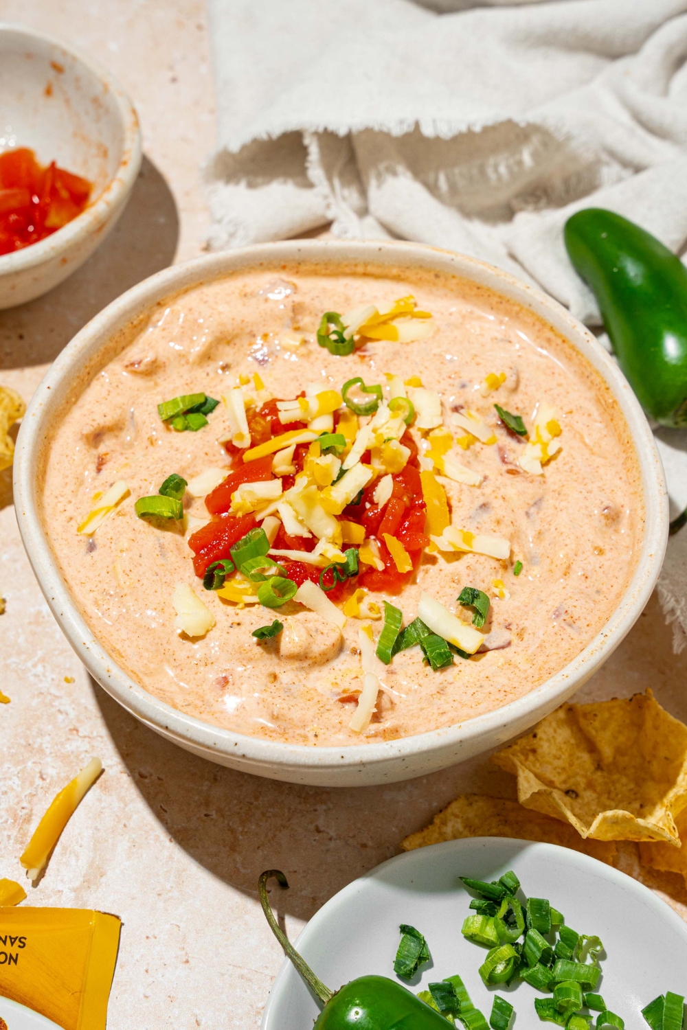 A white bowl with poolside dip garnished with shredded cheese and sliced green onion. The bowl is on a tan counter with small bowls of garnishes and a white cloth napkin.