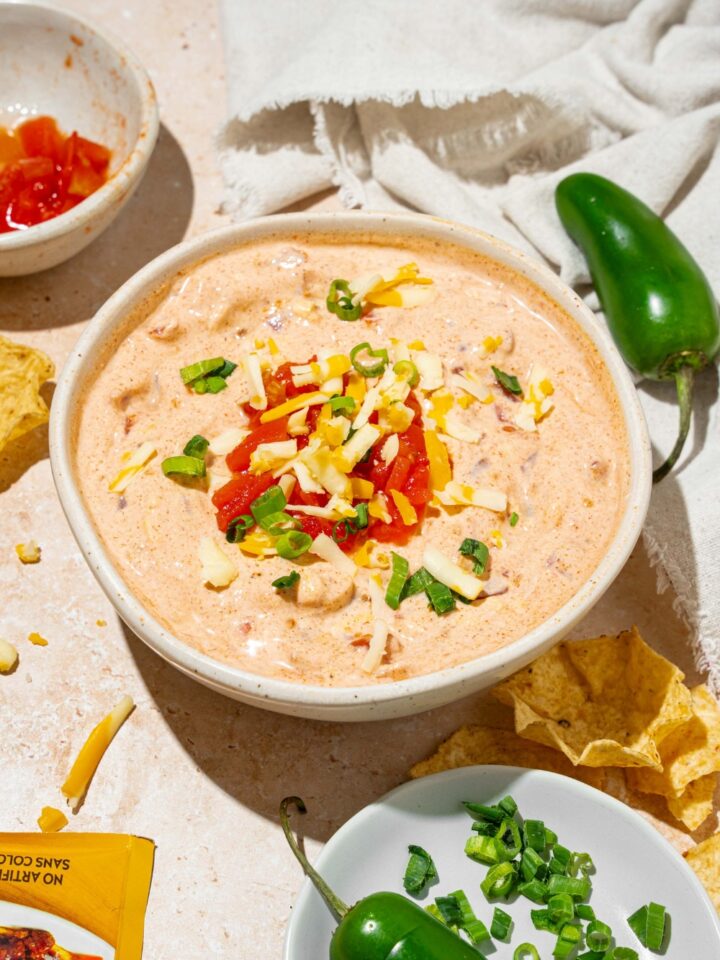 A white bowl with poolside dip garnished with shredded cheese and sliced green onion. The bowl is on a tan counter with small bowls of garnishes and a white cloth napkin.