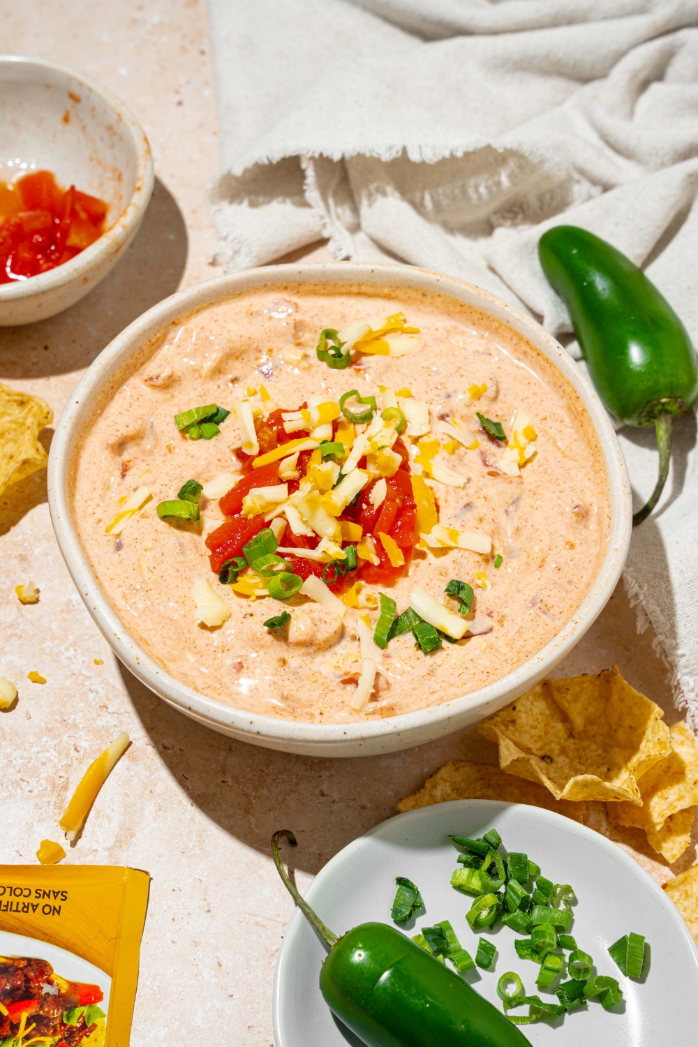 A white bowl with poolside dip garnished with shredded cheese and sliced green onion. The bowl is on a tan counter with small bowls of garnishes and a white cloth napkin.