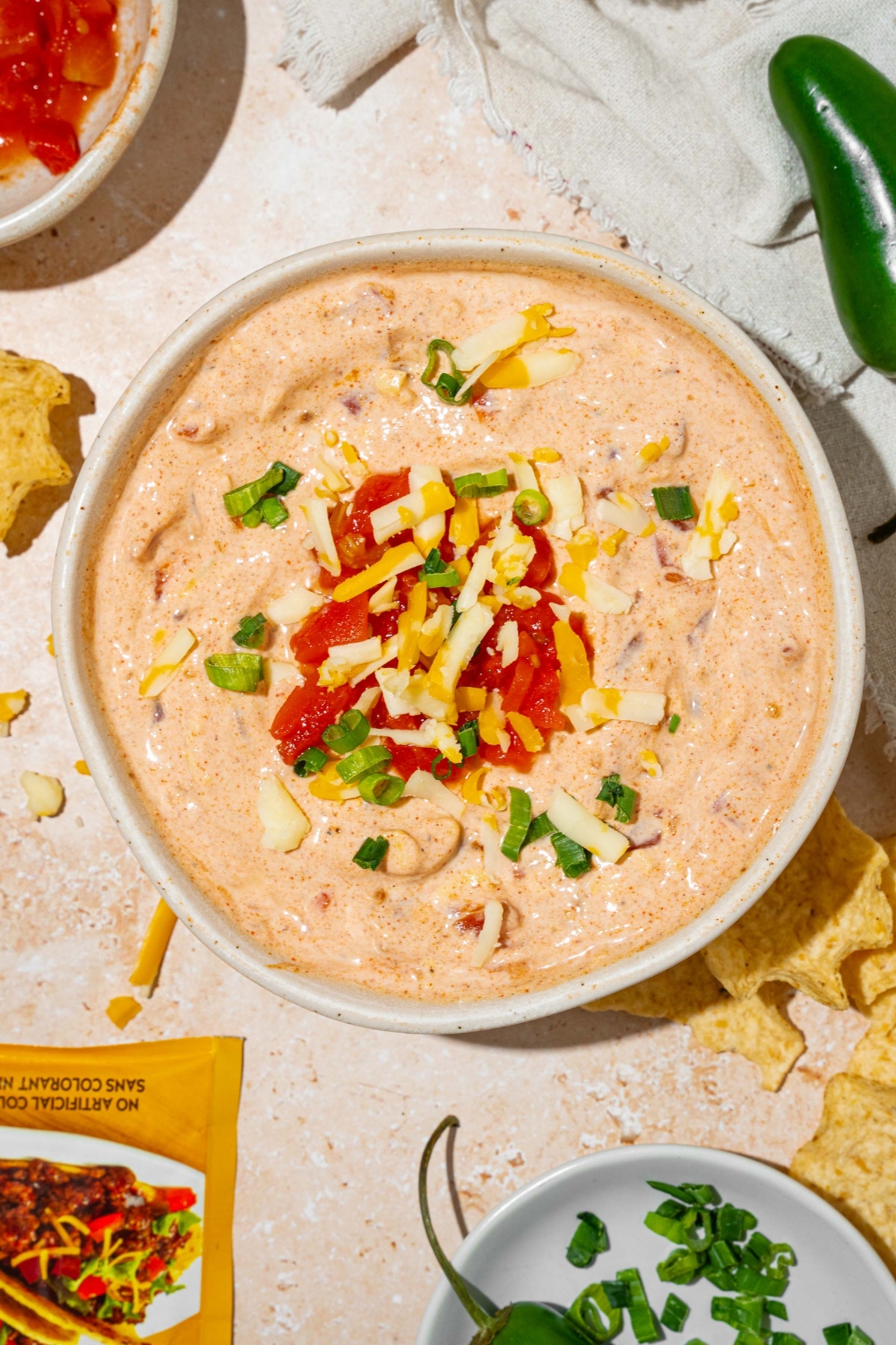 A white bowl with poolside dip garnished with shredded cheese and sliced green onion. The bowl is on a tan counter with small bowls of garnishes and a white cloth napkin.
