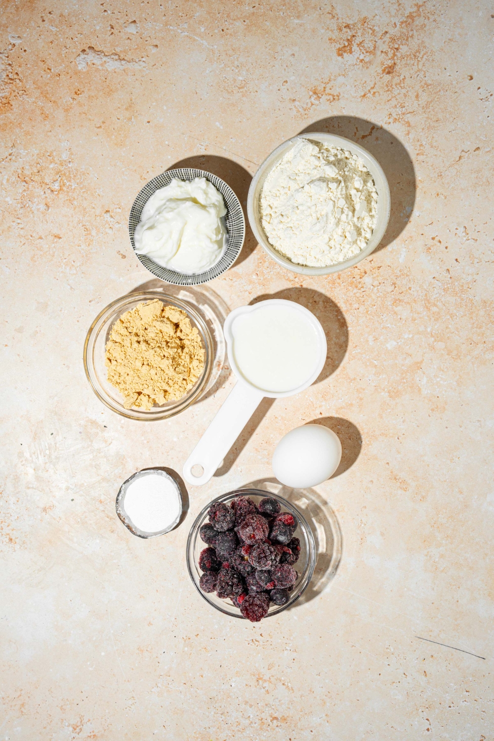 A tan counter with several bowl in various sizes containing ingredients to make protein pancake bowl including flour, yogurt, egg, milk, protein powder, frozen blueberries, and more.