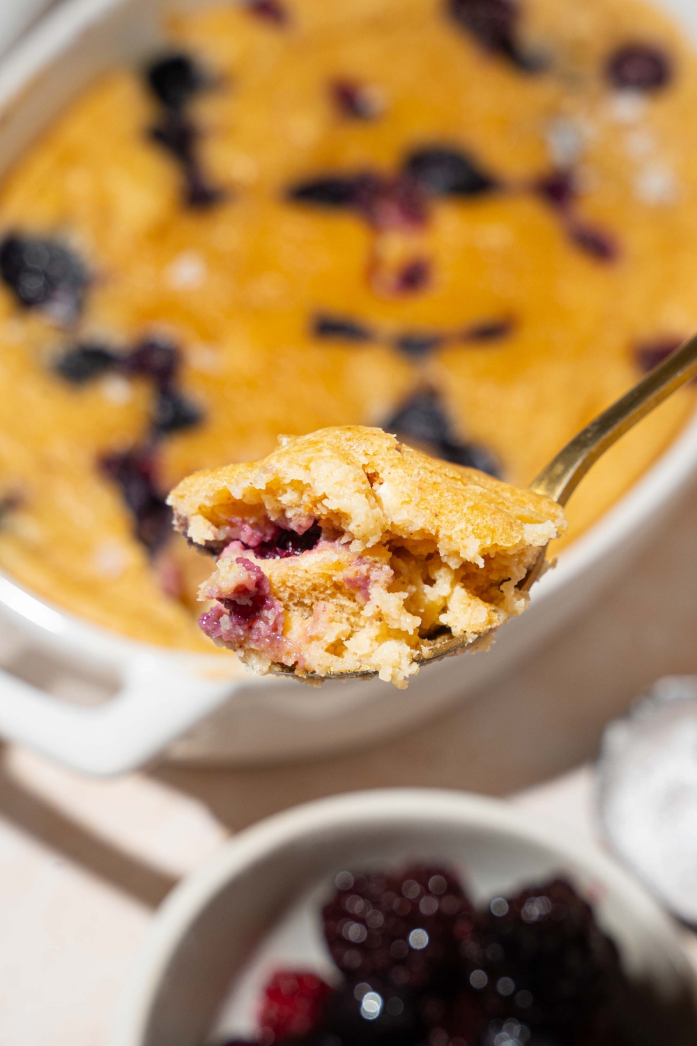 A close up of a spoon with a bite of a protein pancake bowl. The pancake bowl is mixed with blueberries. The bowl is on a tan counter with a small bowl of fruit.