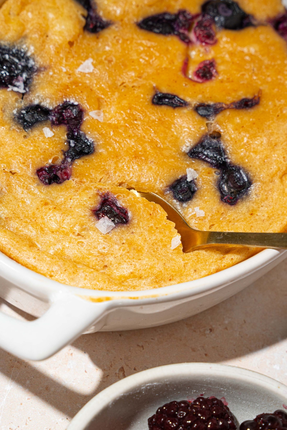 A baked protein pancake bowl with blueberries. There is a spoon taking a bite from the bowl. The bowl is on a tan counter with a small bowl of fruit.