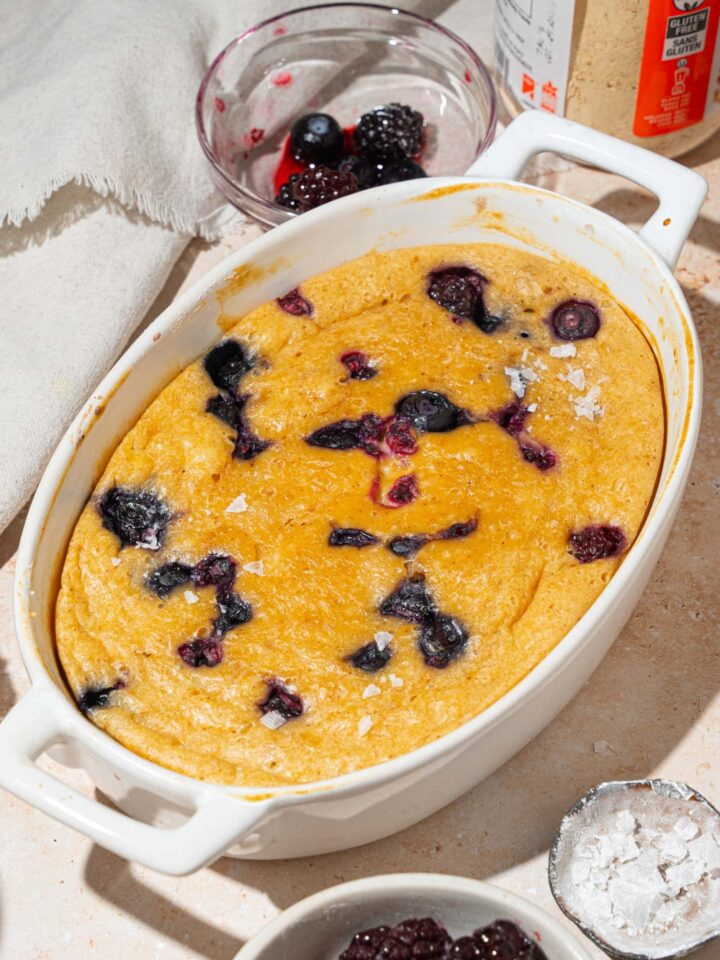 A baked protein pancake bowl with berries. The bowl is on a tan counter with a white cloth napkin and small bowl of fruit.